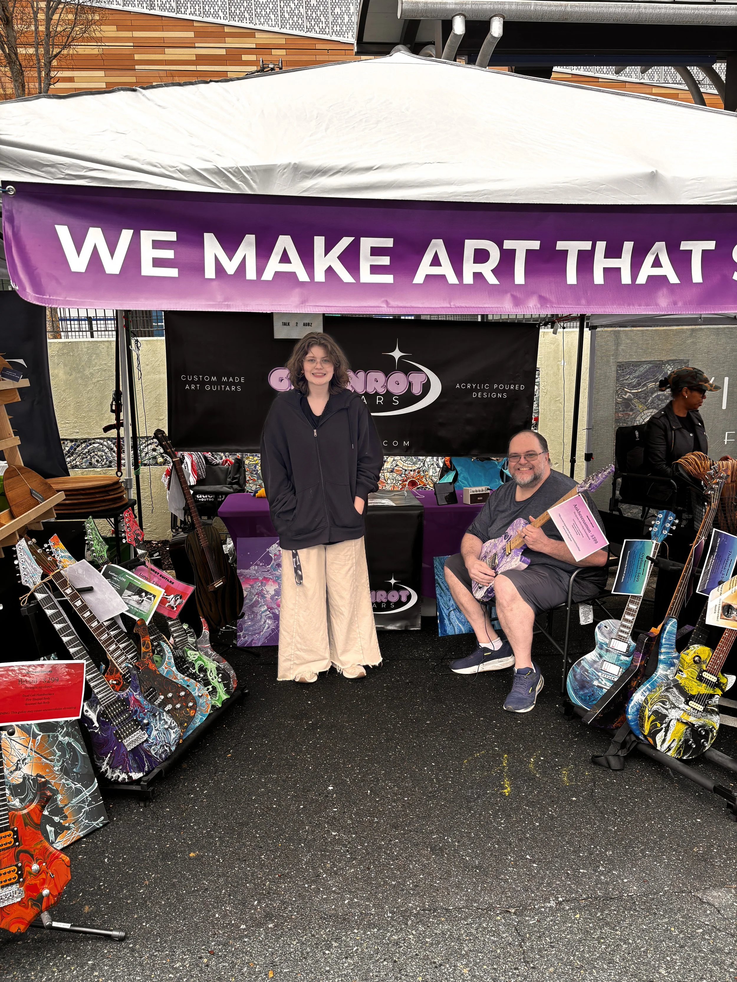 Two people at a booth displaying custom painted guitars, with one person sitting and holding a guitar, and the other standing beside him. The booth has a banner that says, 'WE MAKE ART THAT...' and a black dealer banner behind them. Various colorful guitars are displayed on stands on either side of the booth.