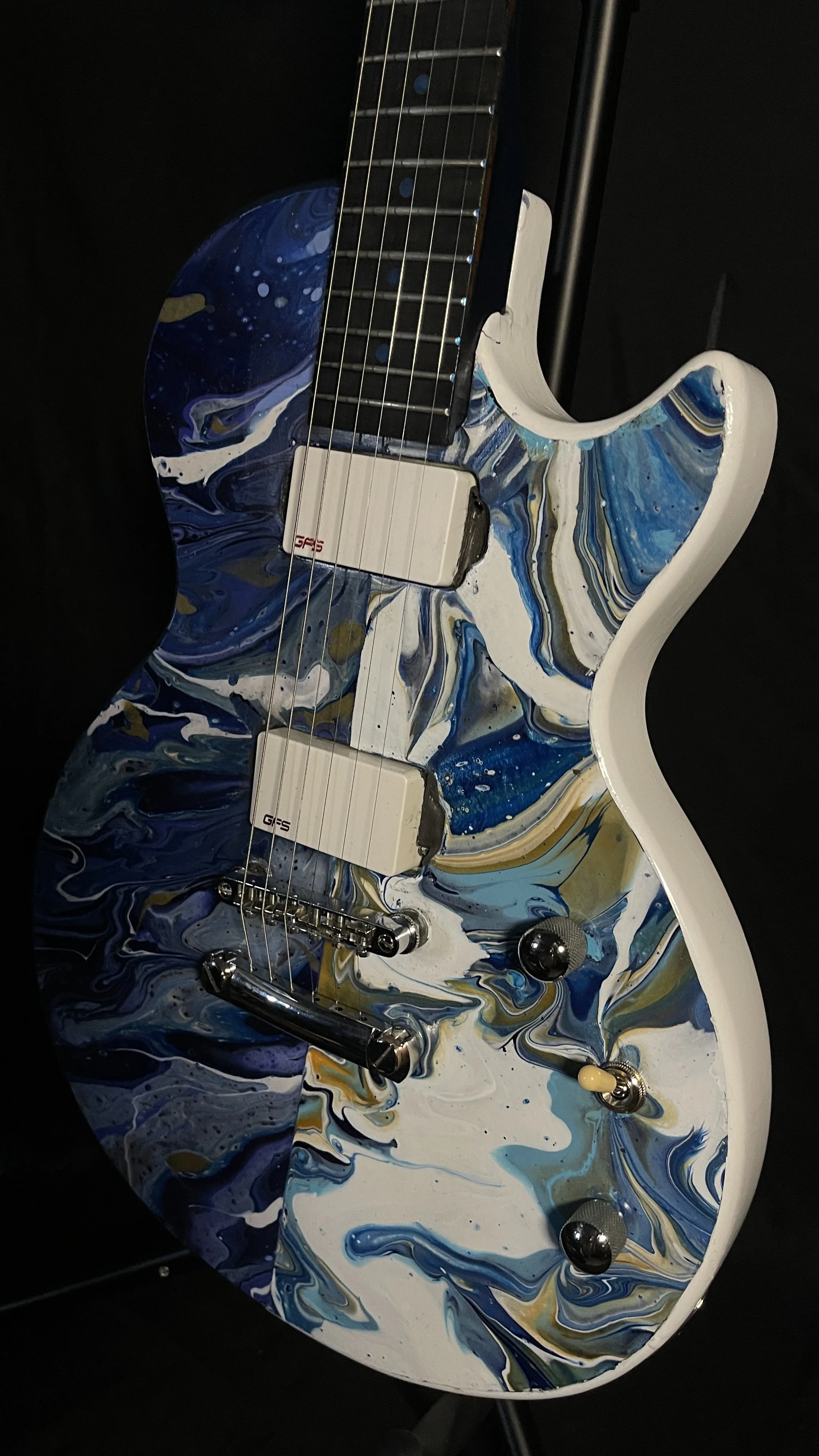 A split acrylic poured electric guitar, one side being dark blue, black and purple, while the other being white, light blue and gold.