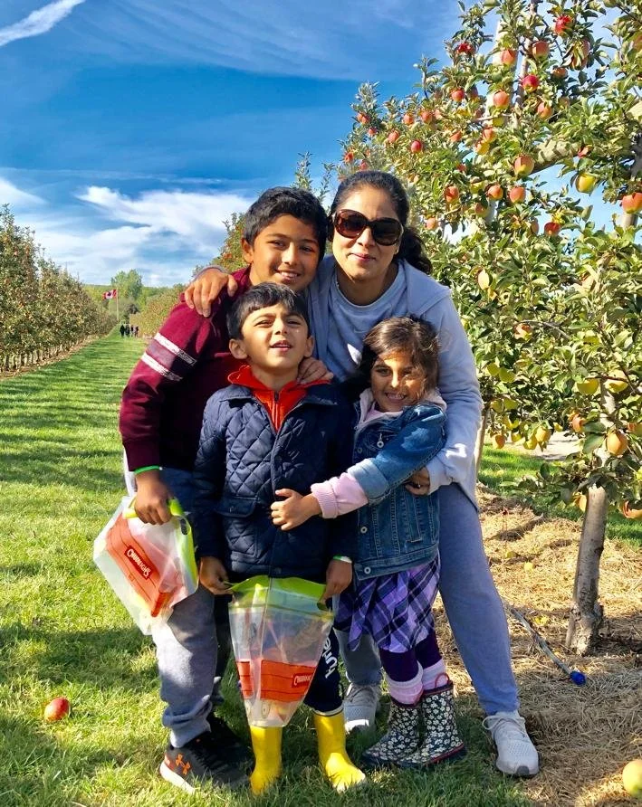 A woman and four children standing in an apple orchard on a sunny day, with apple trees in the background. The woman is wearing sunglasses and a light blue hoodie, and the children are dressed warmly, some holding apple-picking bags.