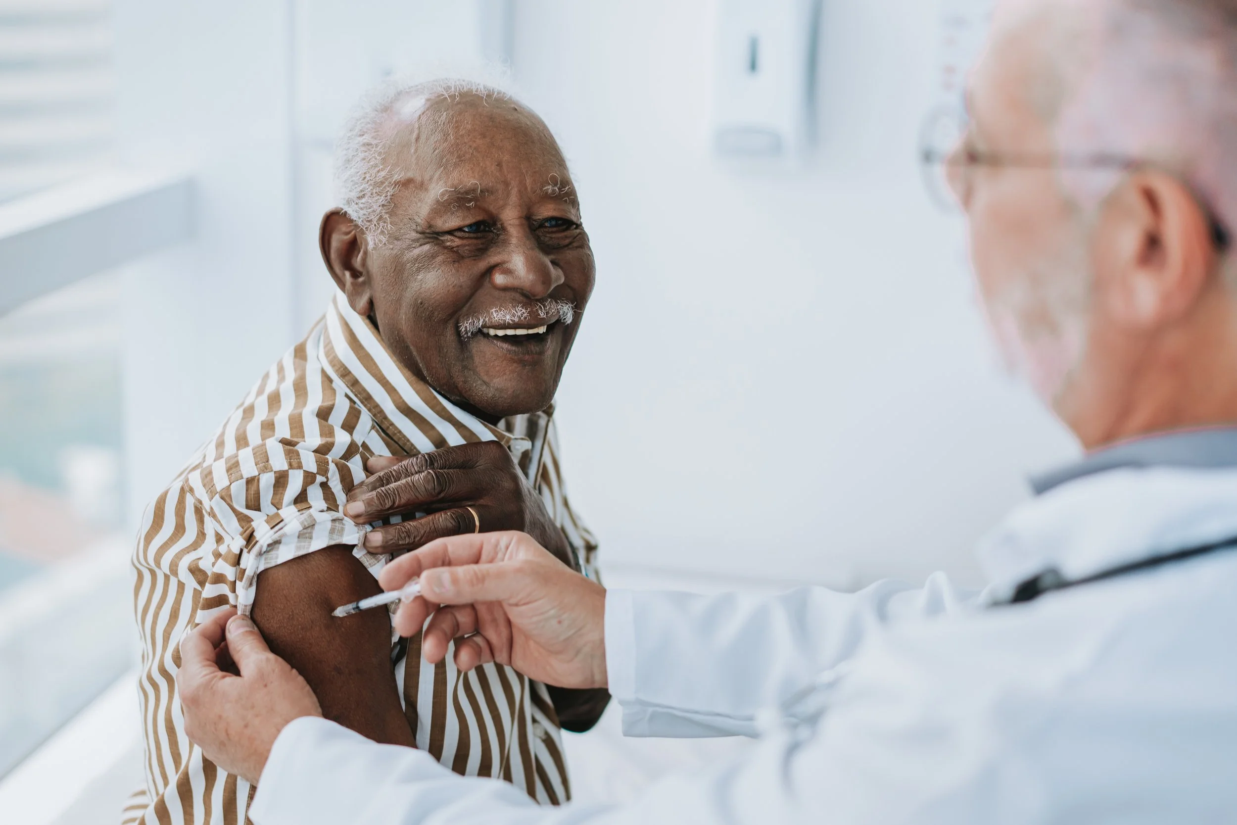 An elderly man is receiving a flu shot or vaccine in his upper arm from a healthcare professional in a clinical setting. The man is smiling and wearing a striped shirt.