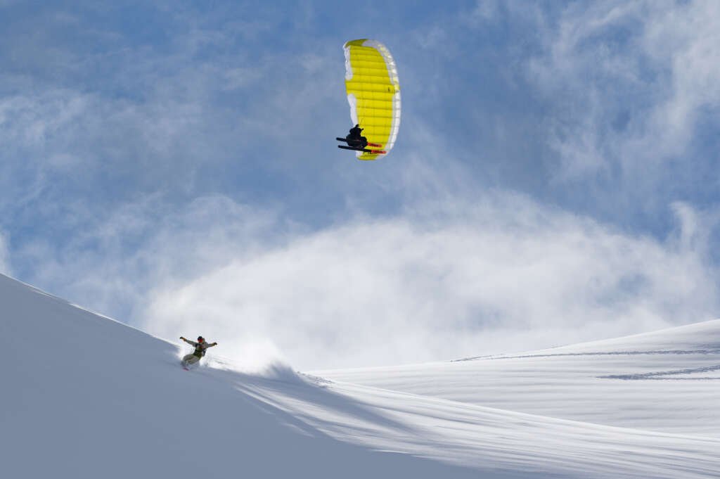 A person snowboarding down a snowy slope with a paraglider flying above in the sky.