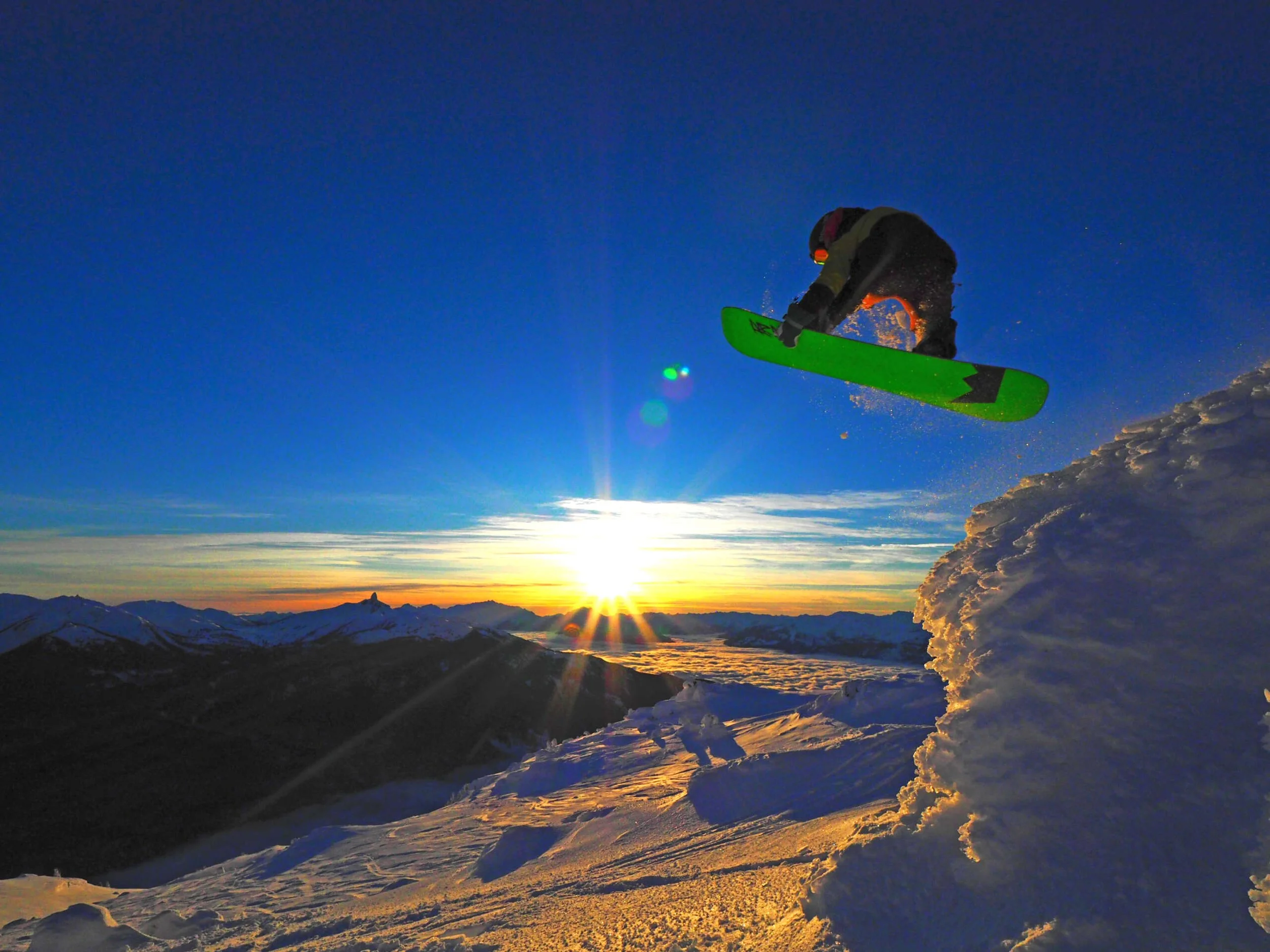 A snowboarder performs a jump off a snowy ledge during sunset over snowy mountains.