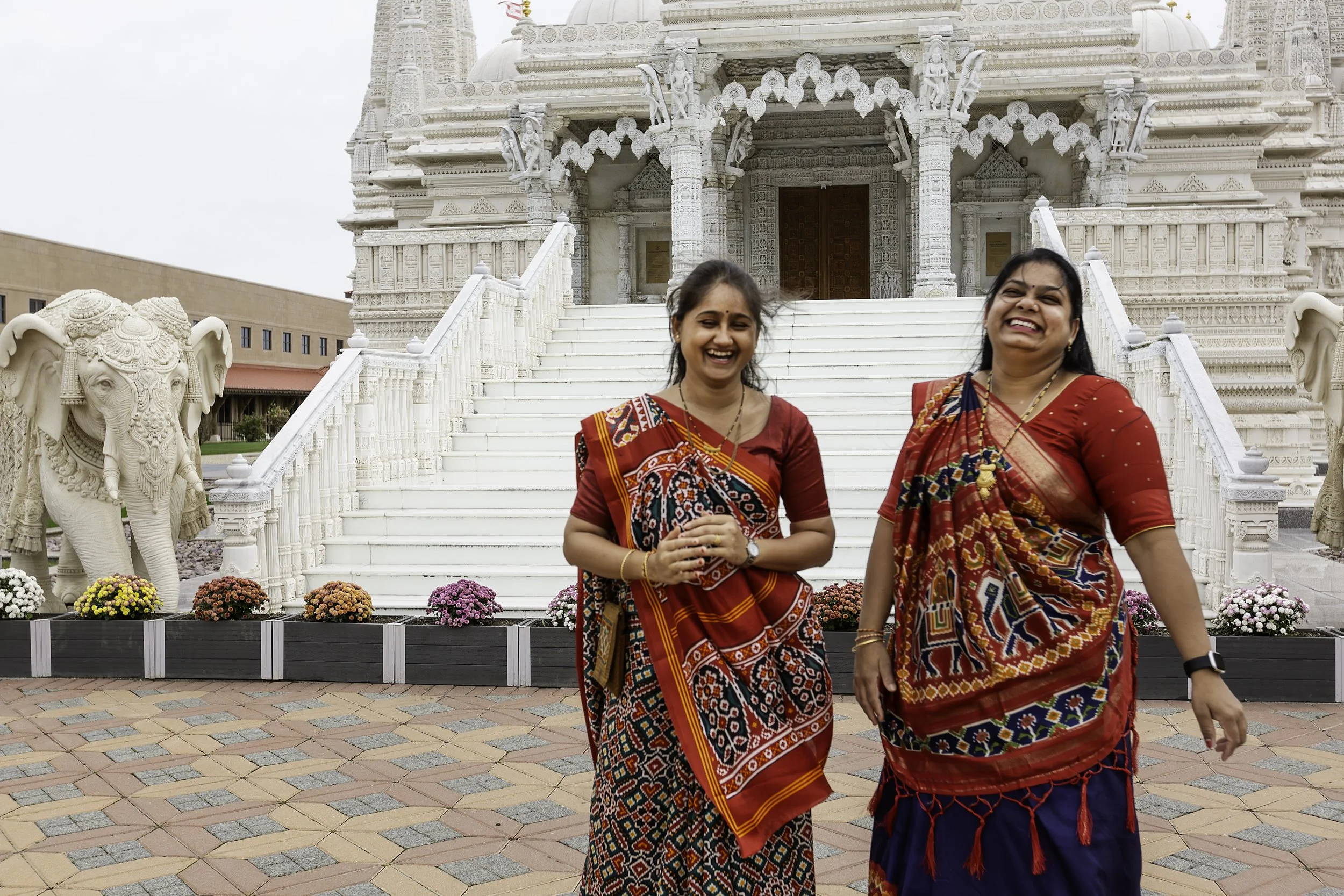 Two women dressed in colorful traditional Indian clothing standing in front of a white temple with intricate carvings, stairs, and sculptures of elephants.