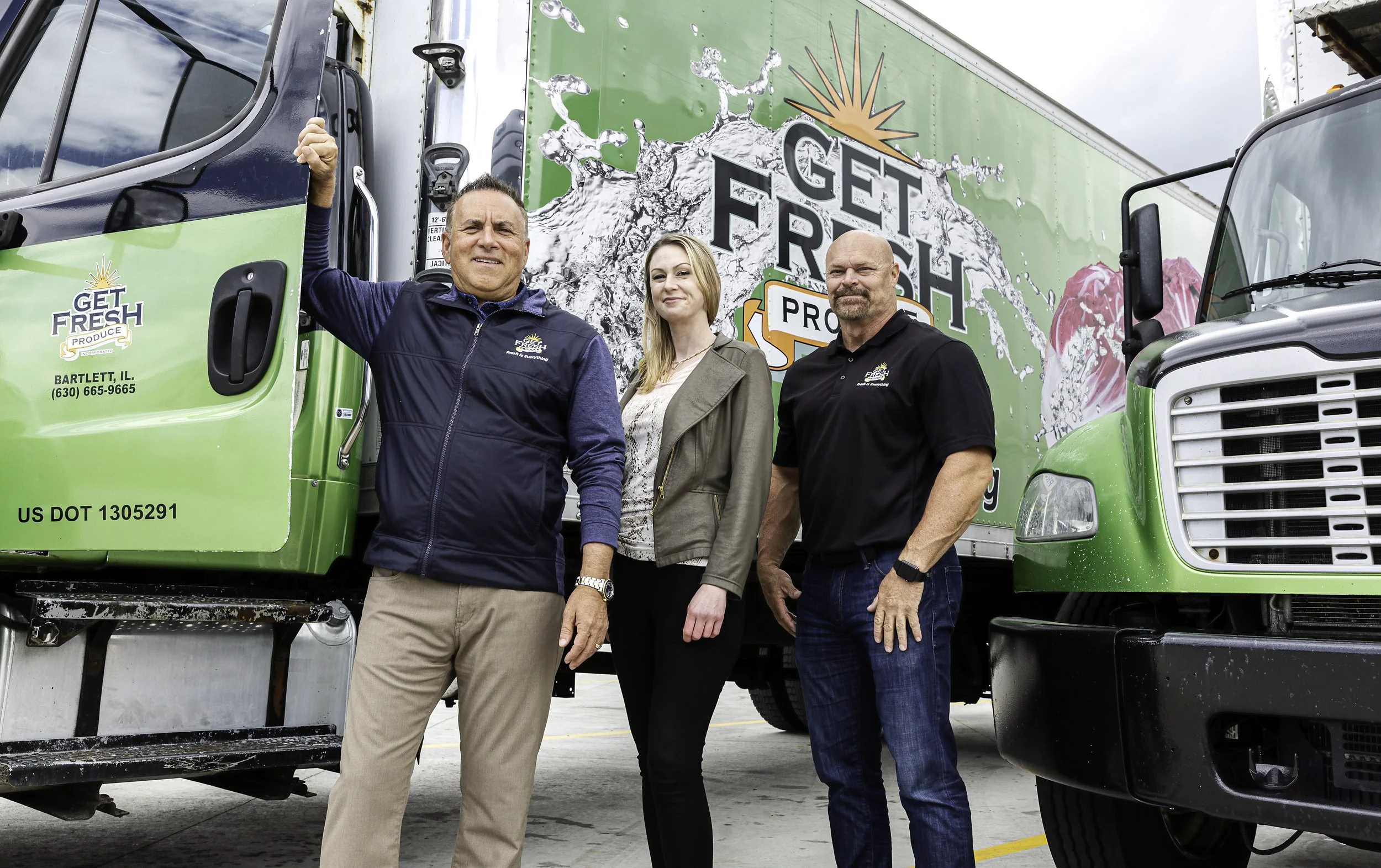 Three people standing in front of a green delivery truck with the logo "Get Fresh Produce" on it. The person on the left is smiling and holding onto the truck door, the woman in the middle is smiling with arms crossed, and the man on the right is looking at the camera with a serious expression.