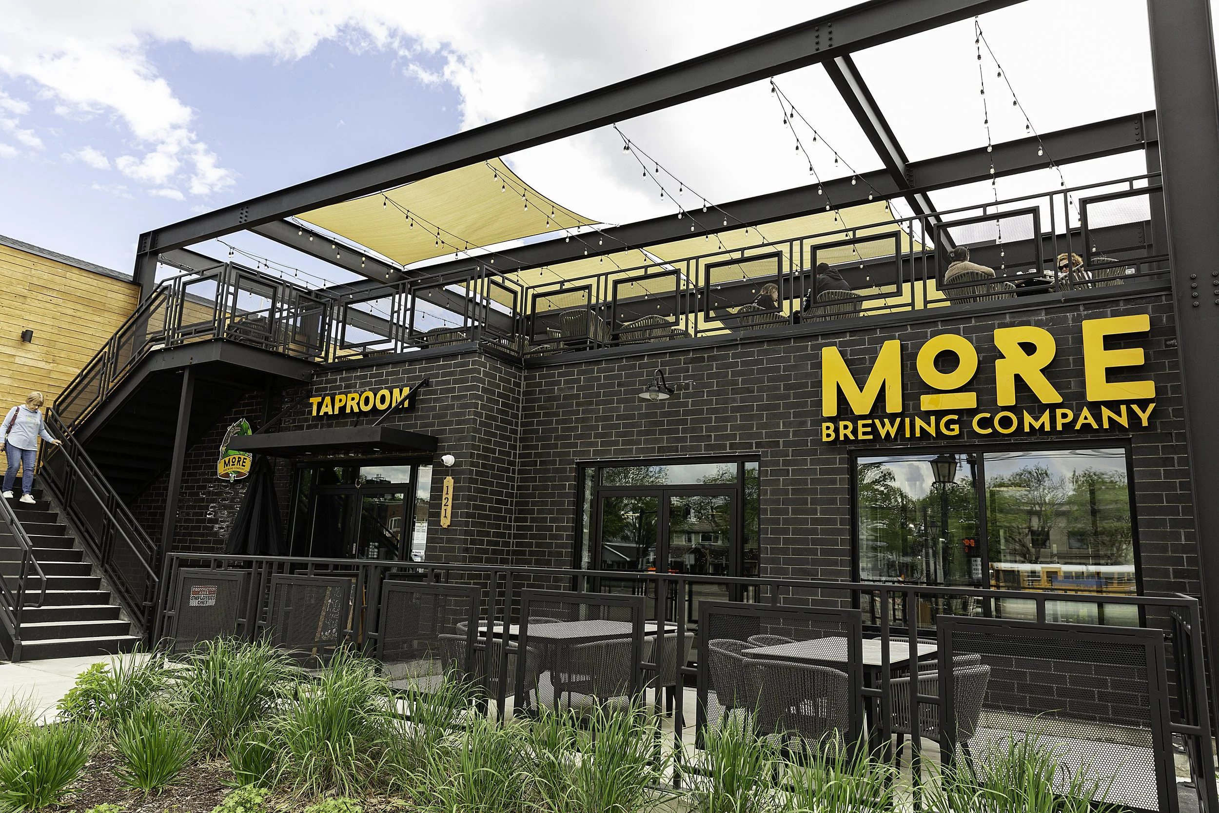 Exterior view of More Brewing Company with outdoor seating, black brick walls, and a rooftop patio with string lights and beige shade sails. A person is walking down stairs on the left.