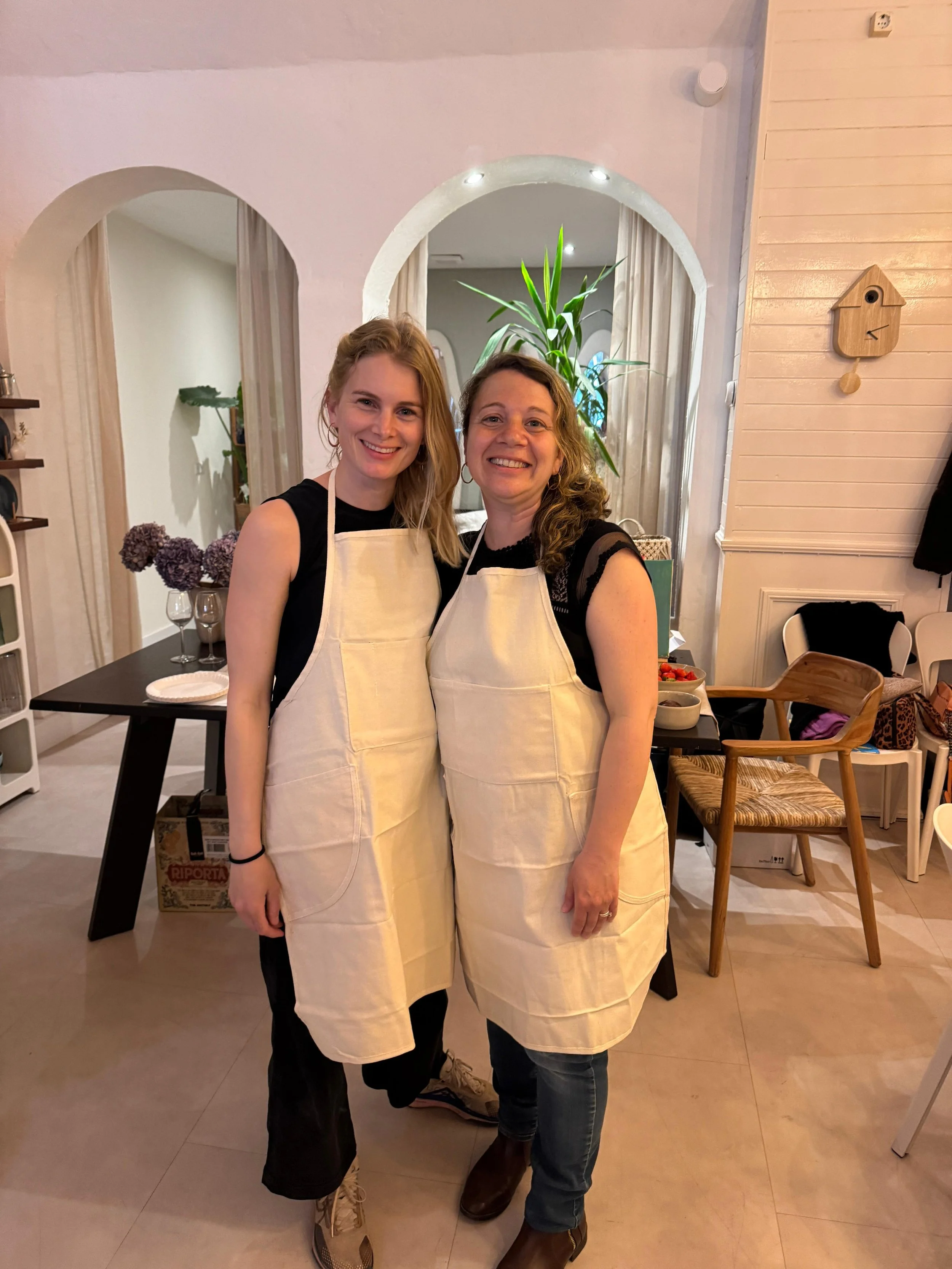 Two women wearing aprons smiling and posing together in a cozy, well-lit indoor space, with dining tables and chairs in the background.