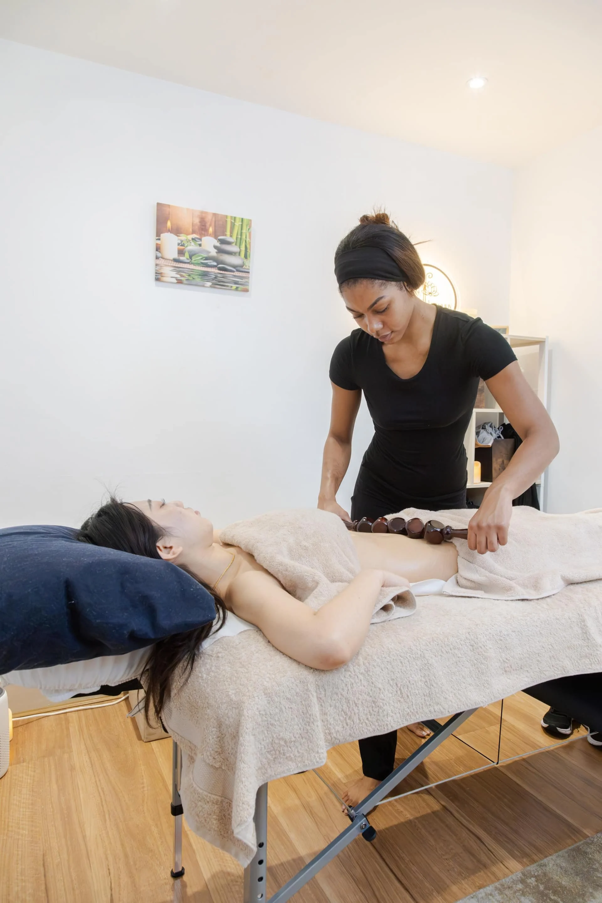 A woman receiving a massage from a masseuse in a spa room, with another woman lying face down on a massage table