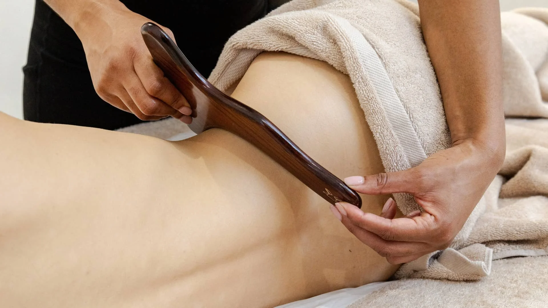 A person in black attire receives a massage using a wooden tool on their thigh, with a beige towel covering parts of their body.