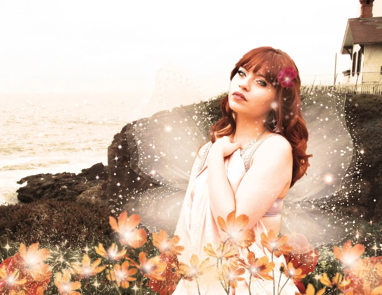 A woman with red hair and fairy wings standing outdoors near a rocky coastline with a house on a hill in the background, surrounded by flowers and sparkles.