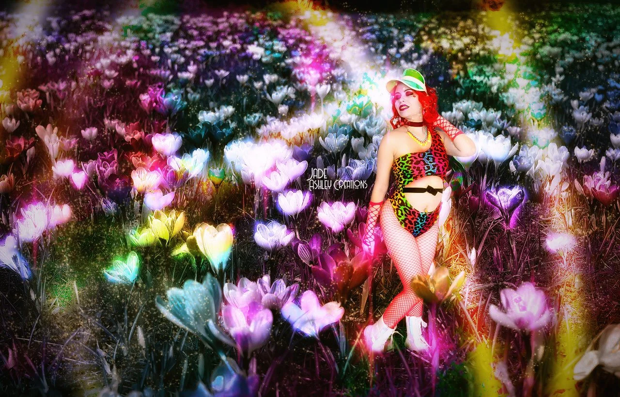 A woman with red curly hair wearing colorful leopard print lingerie, fishnet stockings, a green visor, and jewelry stands among a vibrant field of glowing, multicolored heart-shaped flowers. She poses with one hand behind her head, gazing into the di