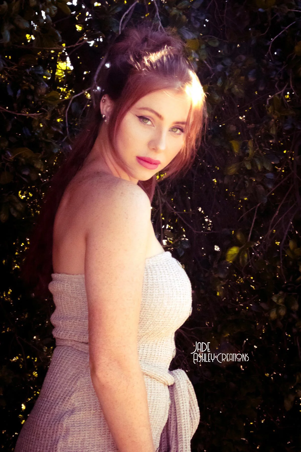 A woman with long reddish-brown hair posing outdoors against dark foliage, wearing a beige sleeveless top and minimal makeup.