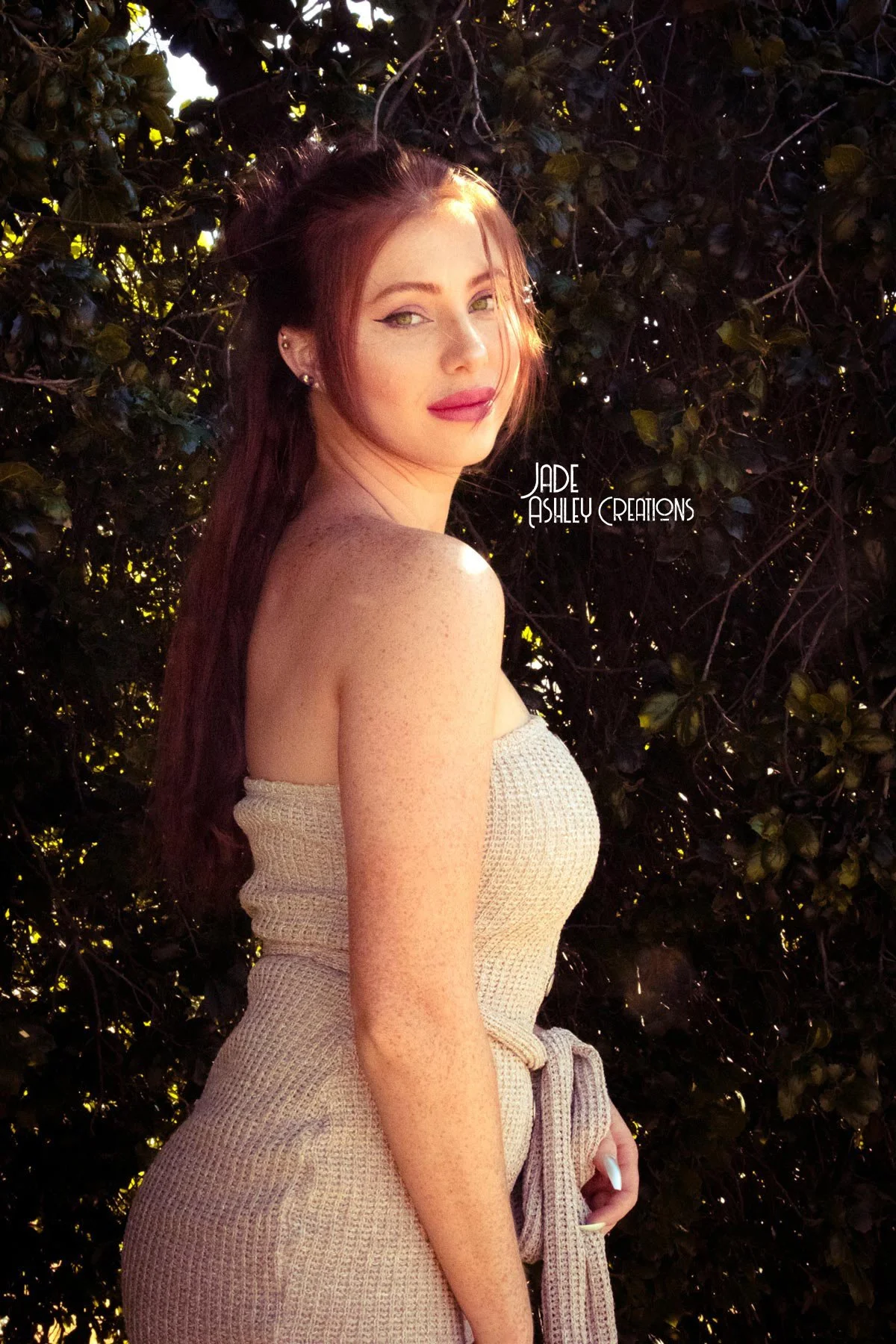 A woman with long reddish-brown hair standing outdoors in front of dense dark green foliage. She is wearing a strapless beige top and matching high-waisted pants, posing with her body turned slightly to the side and looking at the camera. The text 'J