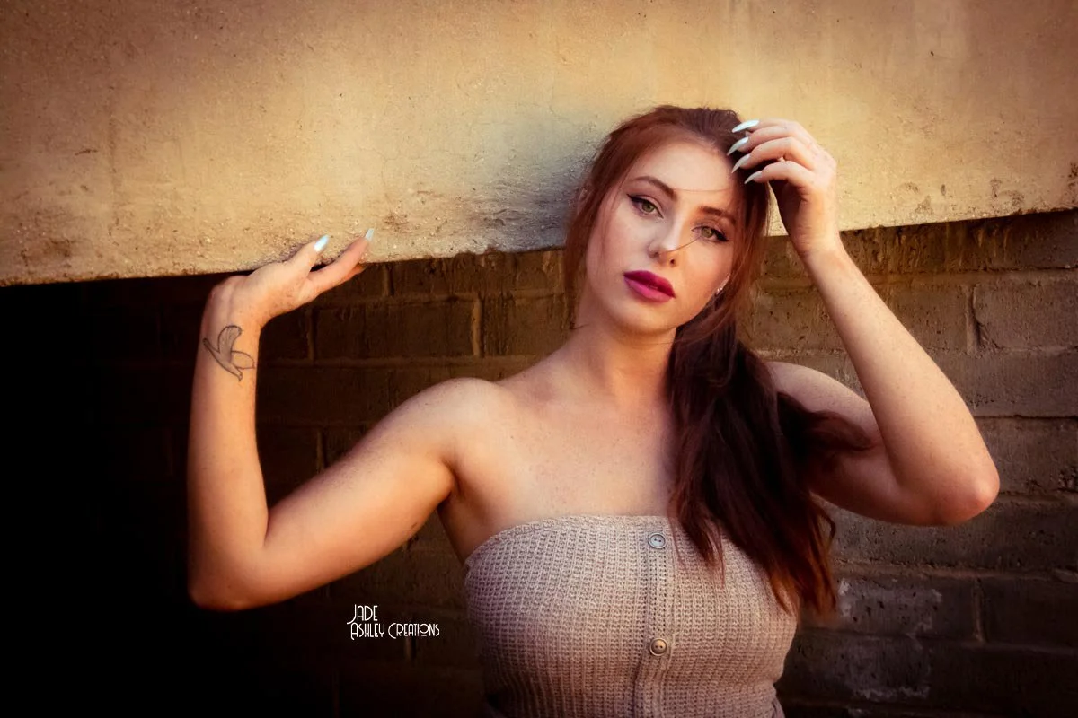 A young woman with long red hair, wearing a strapless beige top, is posing against a brick wall, holding her hair with one hand and touching her forehead with the other.