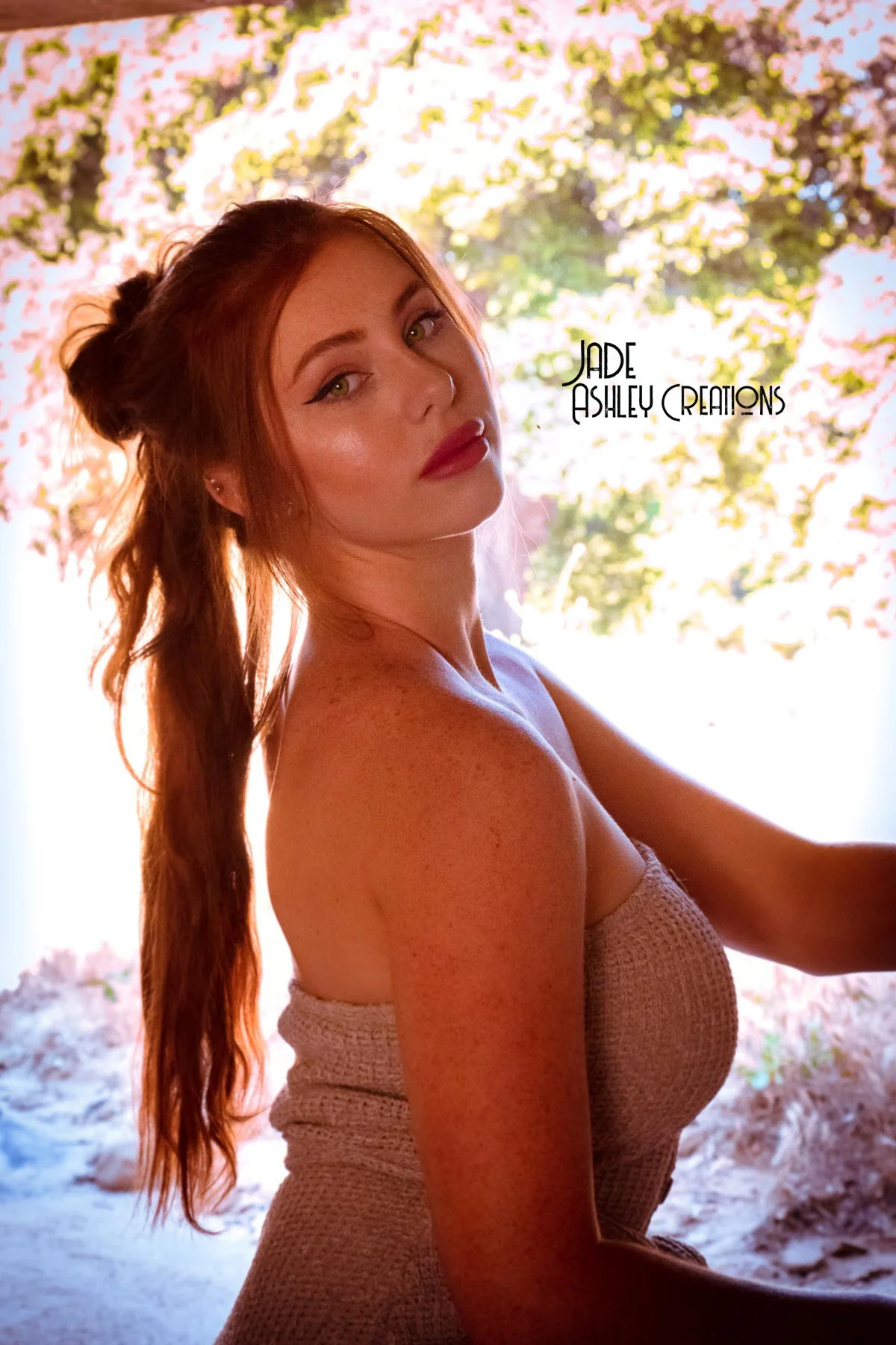 A woman with long red hair and freckles posing outdoors with a blurred background of trees and sunlight.