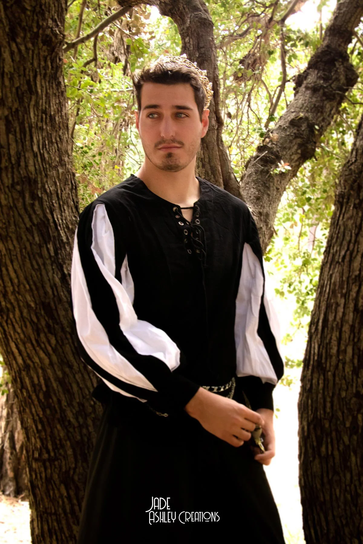 A young man standing between two trees in a forest, wearing a black and white historical costume with a tiara, holding a small object in his hand, with a serious expression.