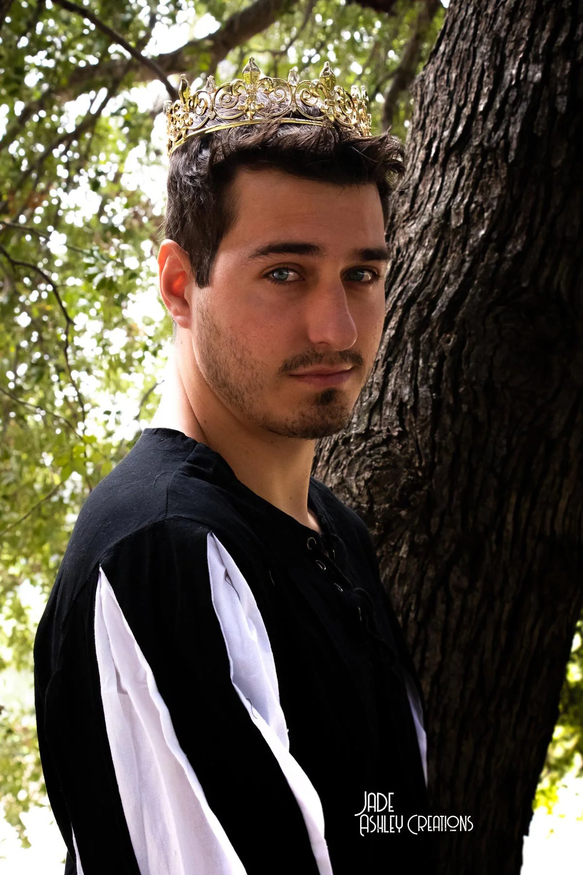 A young man with short dark hair and facial hair, wearing a crown and a black shirt with white stripes, standing outdoors beside a large tree with green leaves in the background.