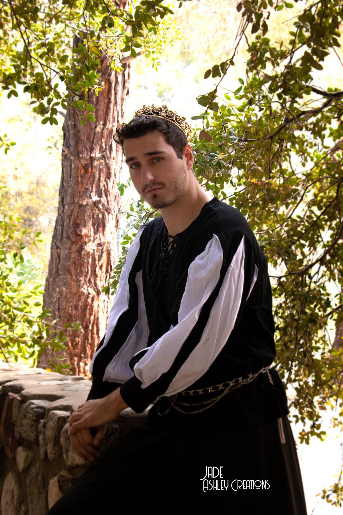 A young man with dark hair and facial hair, wearing a black and white Renaissance-style shirt and a decorative chain belt, poses outdoors against a stone wall. He has a golden crown or circlet on his head and is surrounded by lush green trees and fol