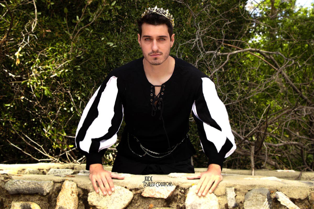 A young man with dark hair and a mustache, wearing a black shirt with white puffed sleeves and a decorative chain, poses outdoors behind a stone wall. He is wearing a small tiara and has a serious expression, with dense green foliage in the backgroun