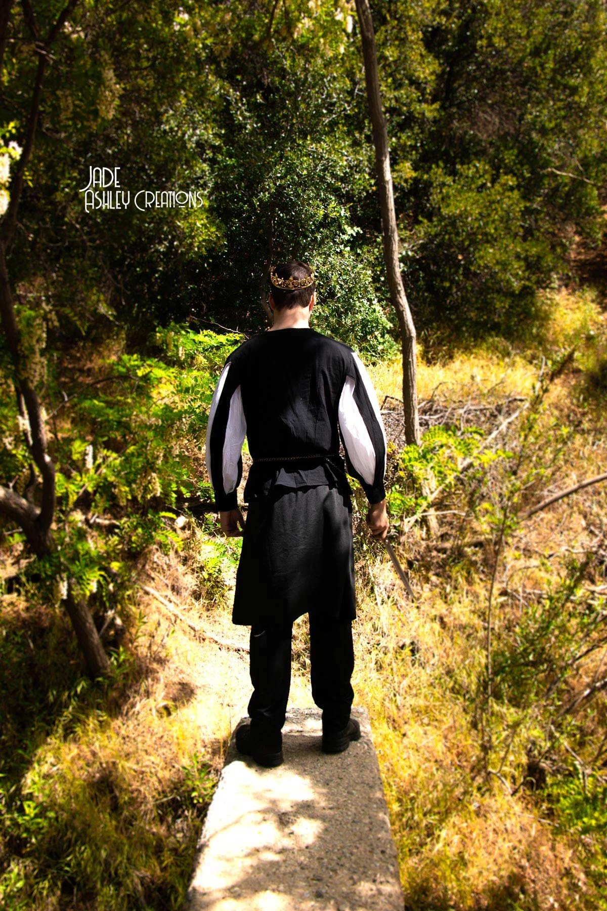 Person wearing a black and white medieval-style outfit with a gold crown, holding a sword, standing on a narrow concrete pathway in a lush wooded area.