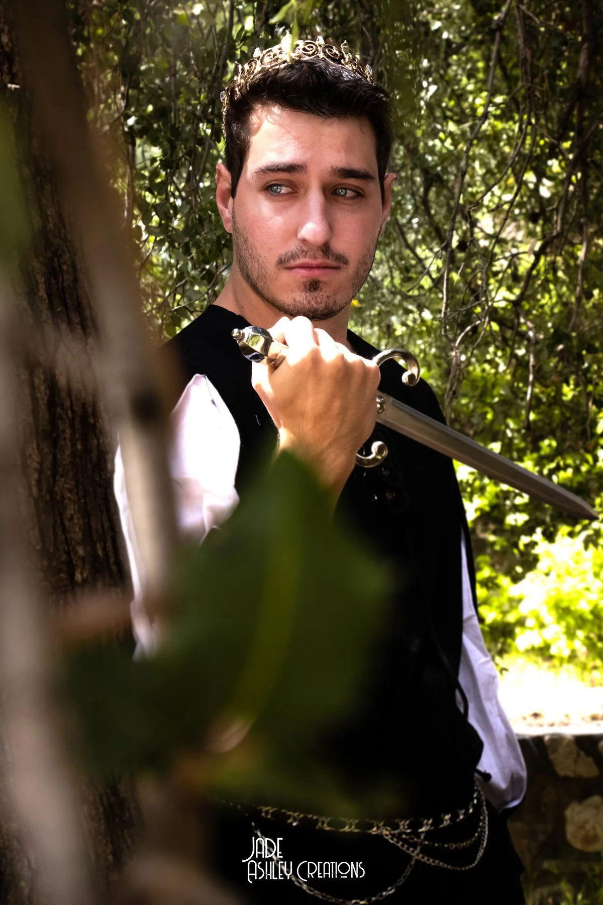 A young man in medieval or fantasy-style costume holding a sword in a wooded area. He wears a chain crown and has a serious expression.