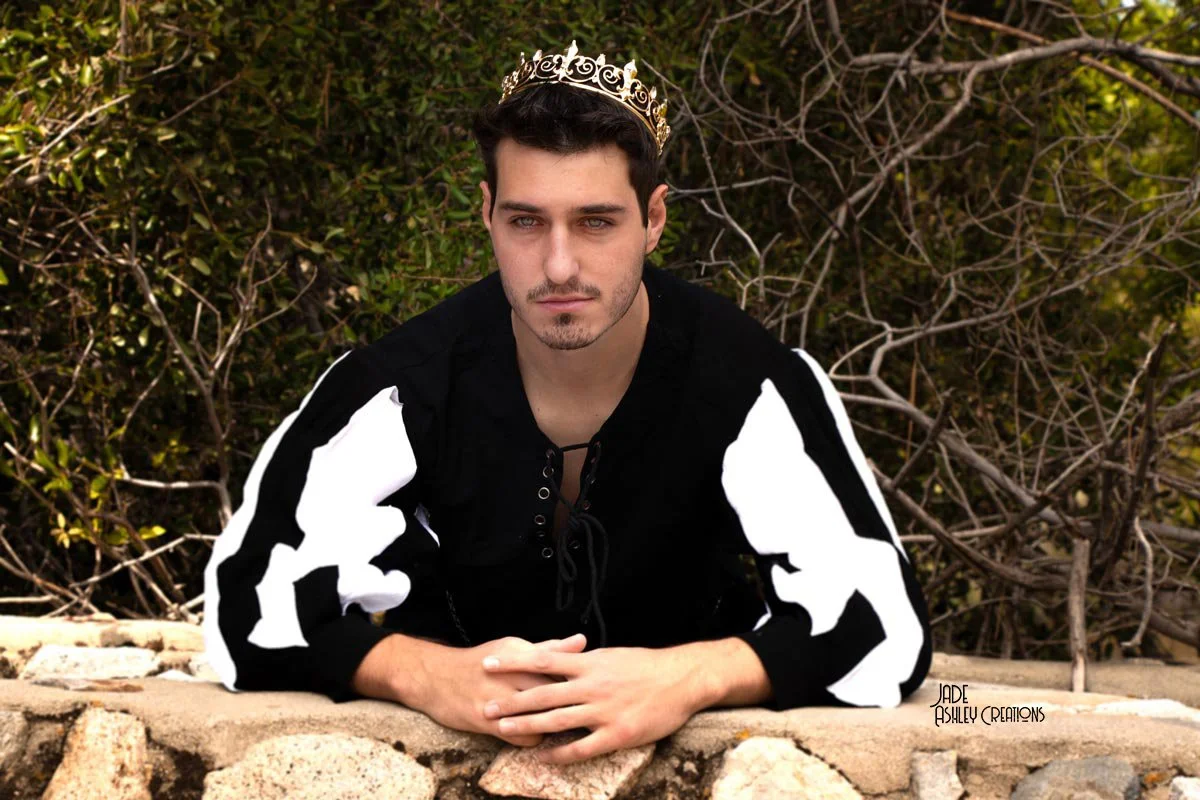 Young man with dark hair and facial hair wearing a black jacket with white sleeves and a gold crown posing outdoors in front of bushes and branches.