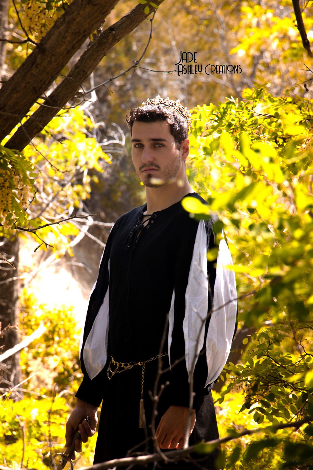 A young man standing outdoors among autumn-colored leaves, wearing a black and white medieval-inspired outfit with a crown and chain accessory, holding a sword, and looking at the camera.