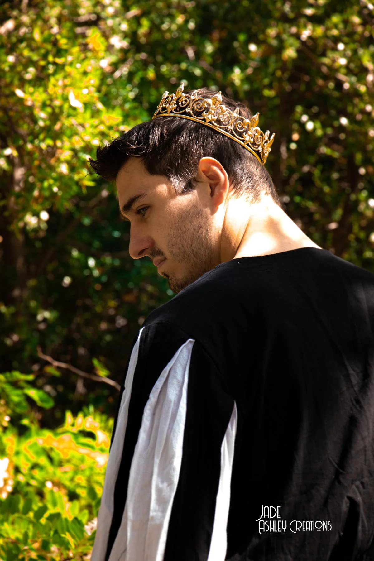 A young man with dark hair and facial hair wearing a golden crown stands outdoors against a backdrop of green foliage. He is looking down and to the left, dressed in a black shirt with white stripes on the sleeves. The photo has natural sunlight and 