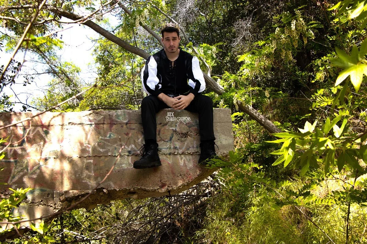 A young man sitting on a graffiti-covered concrete ledge in a forested area surrounded by green trees and foliage.