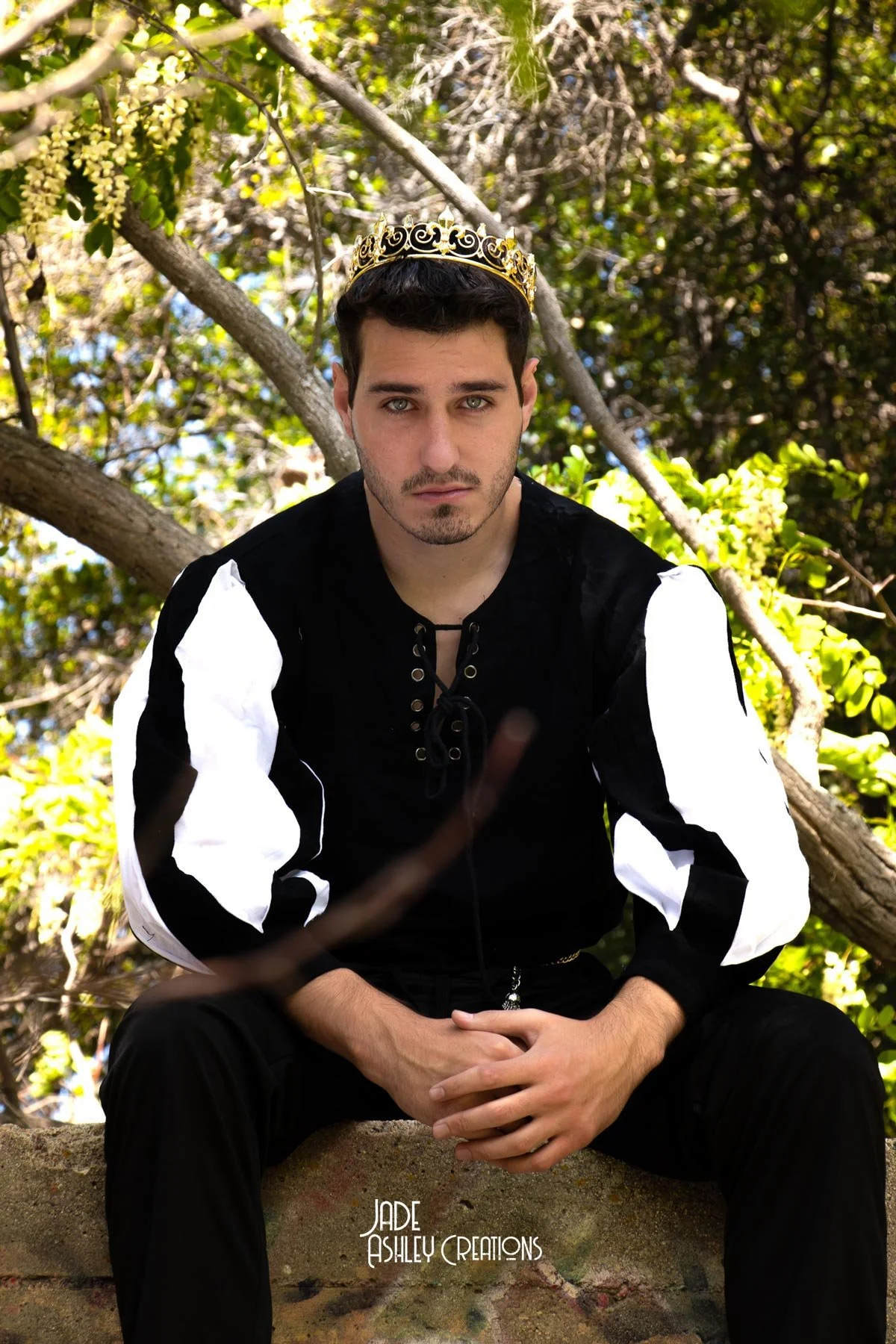 A young man sitting outdoors among trees, wearing a black and white shirt and a gold crown, looking directly at the camera with a serious expression.