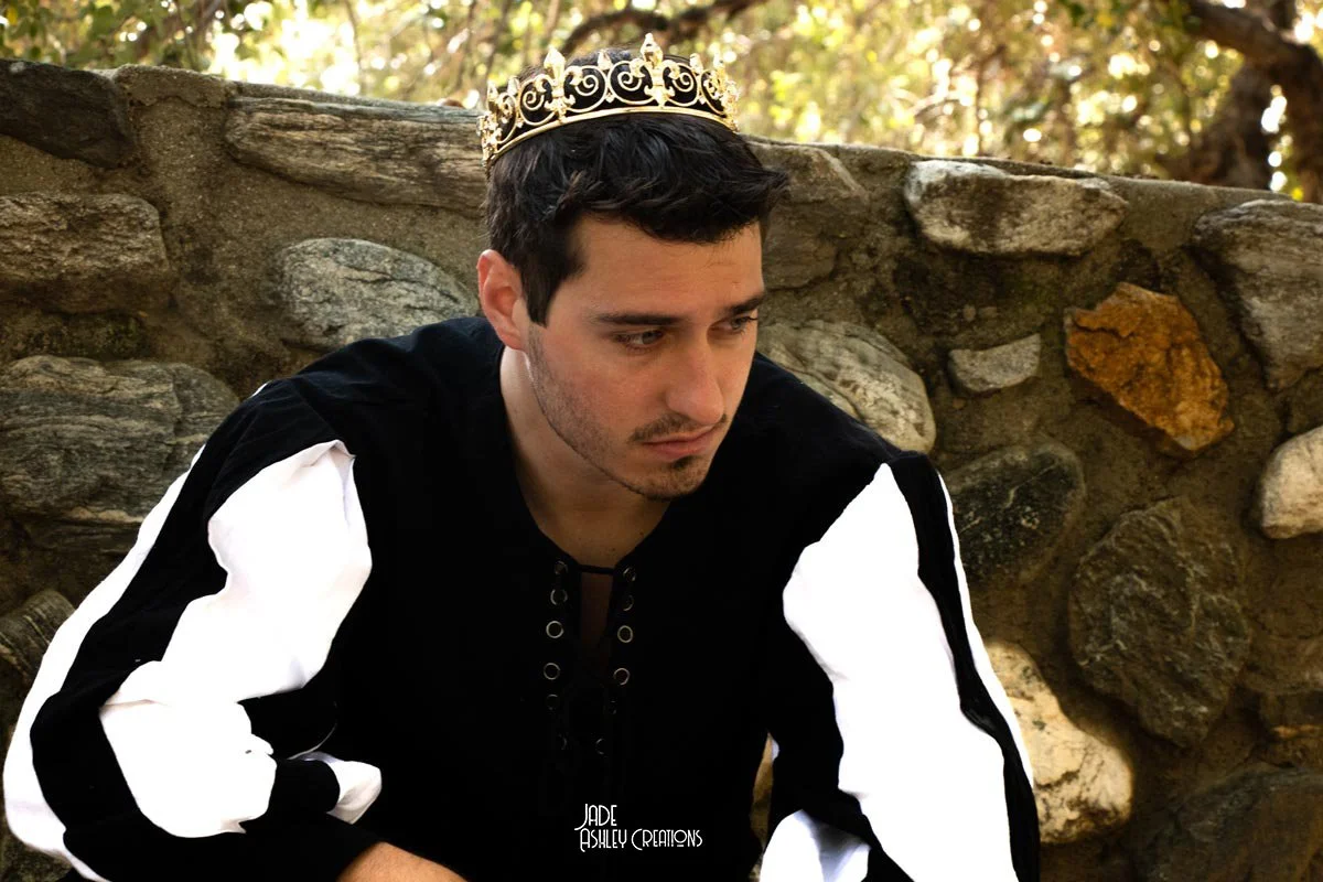 A young man wearing a crown and hoodie, sitting in front of a stone wall outdoors.