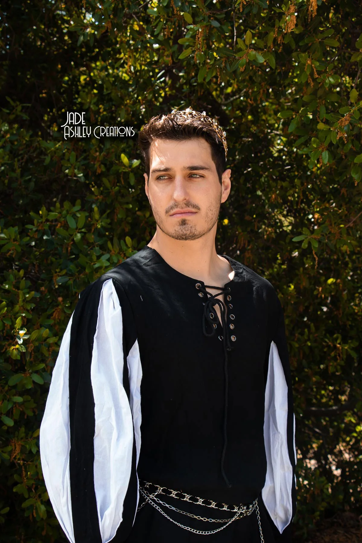 A young man with dark hair and facial hair posing outdoors in front of green foliage, wearing a black and white medieval-inspired shirt with laces and chains.