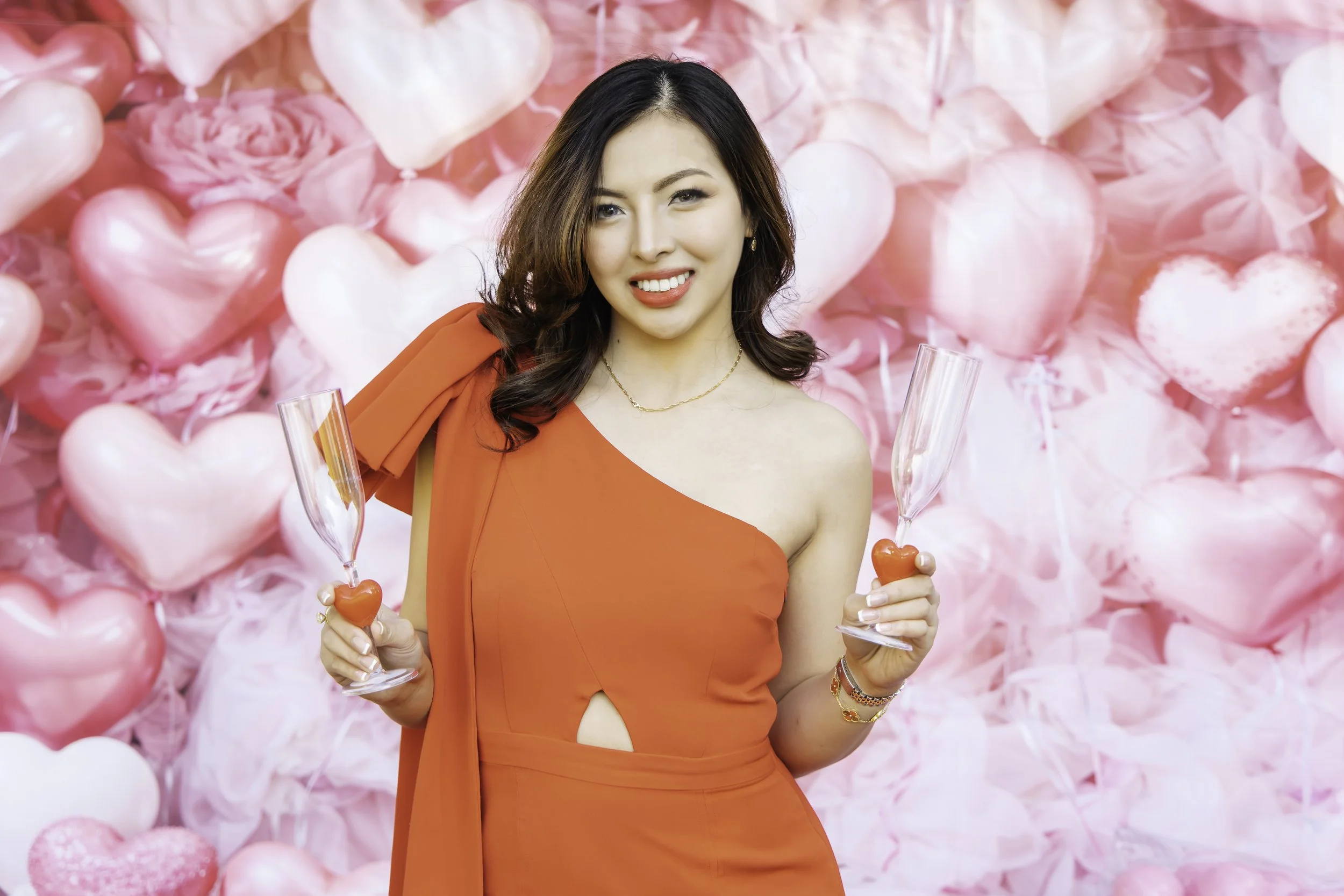 A woman in a red dress holding two champagne glasses with heart-shaped decorations, standing in front of a pink heart-themed backdrop.