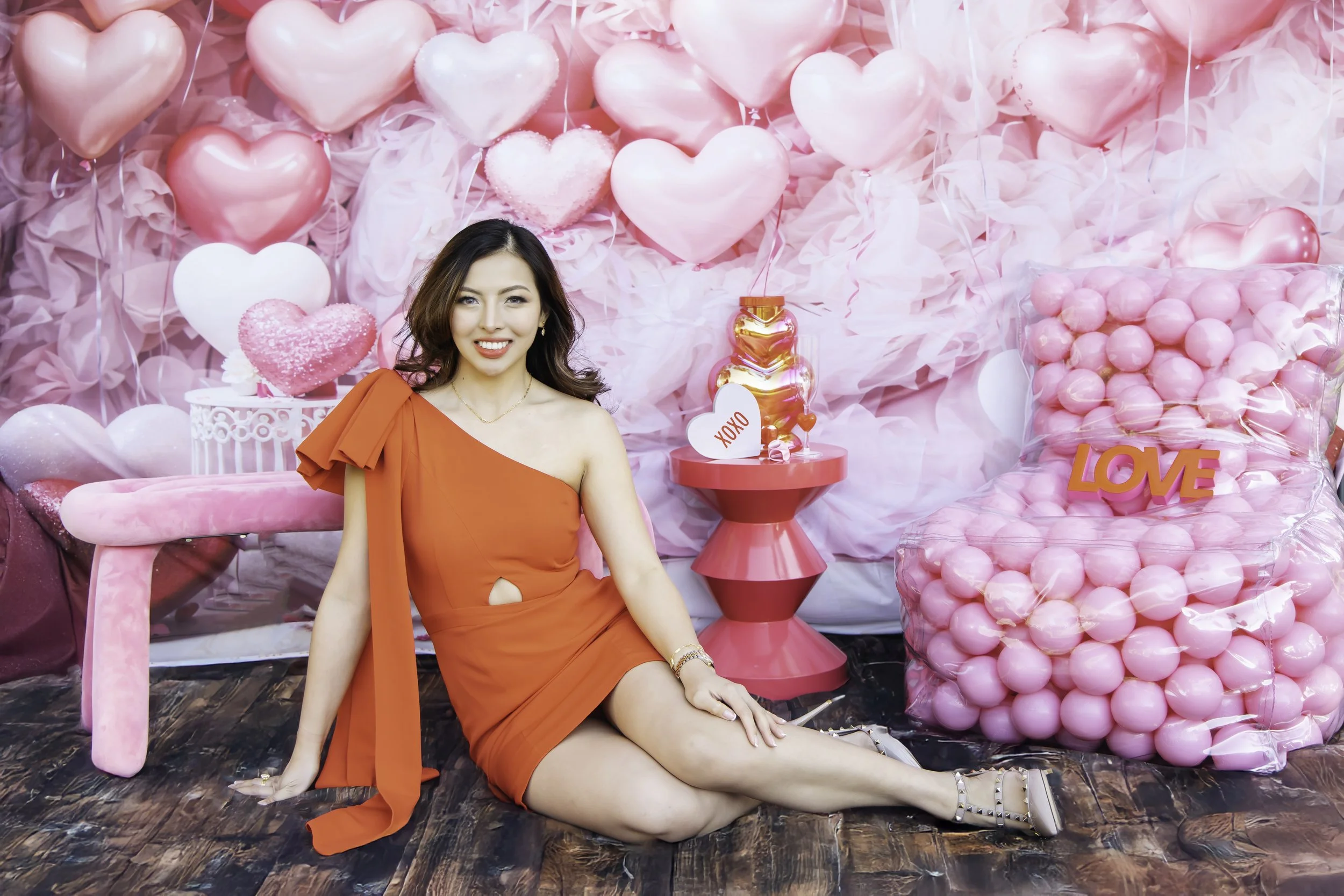 A woman in a red dress sitting on a wooden floor in front of a pink Valentine's themed backdrop with heart-shaped balloons, a pink table, and a bag of pink balls with the word 'LOVE'. She is smiling and posed with one leg extended and the other bent.