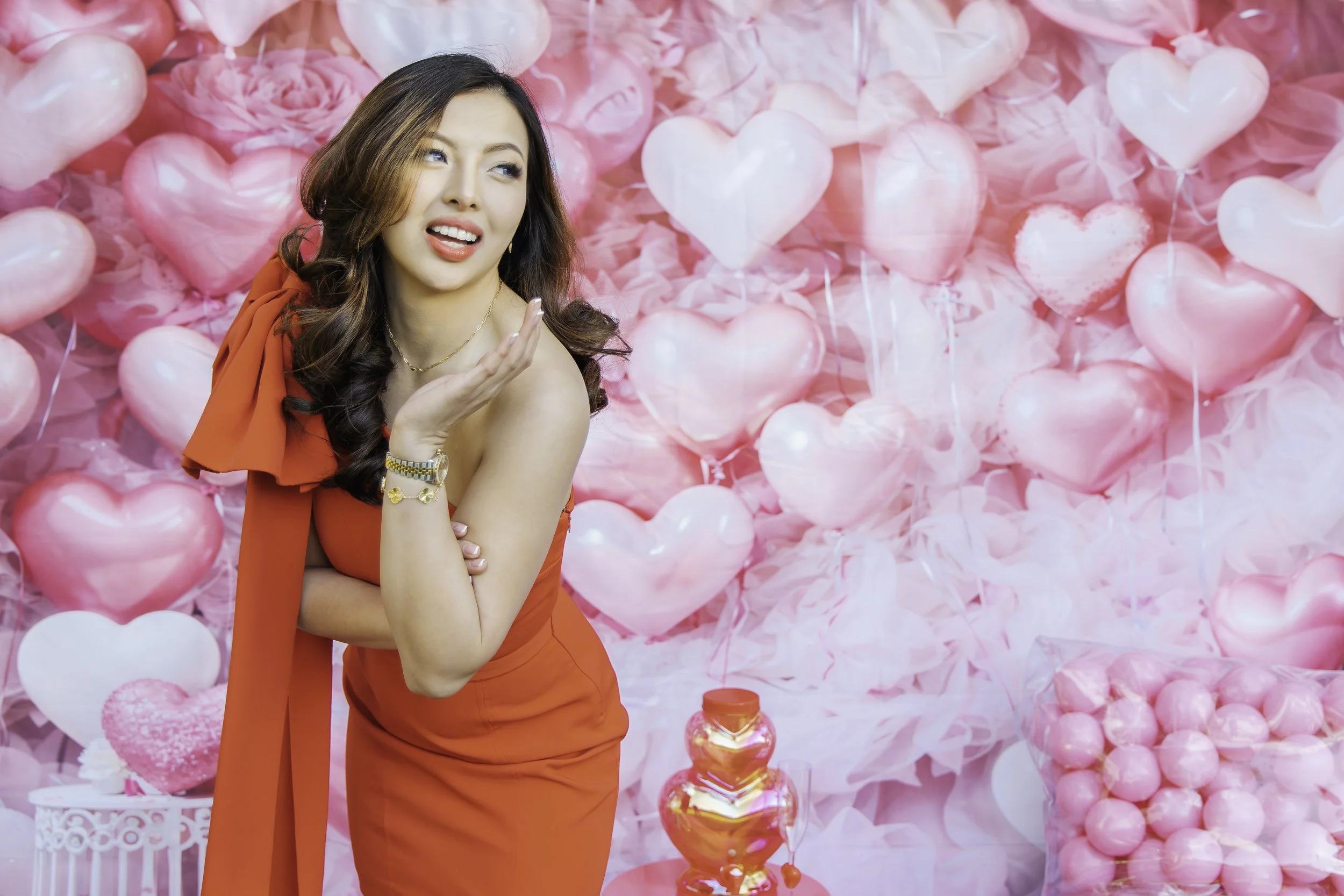 A woman in a red dress posing against a heart-themed pink background with balloons and decorations.
