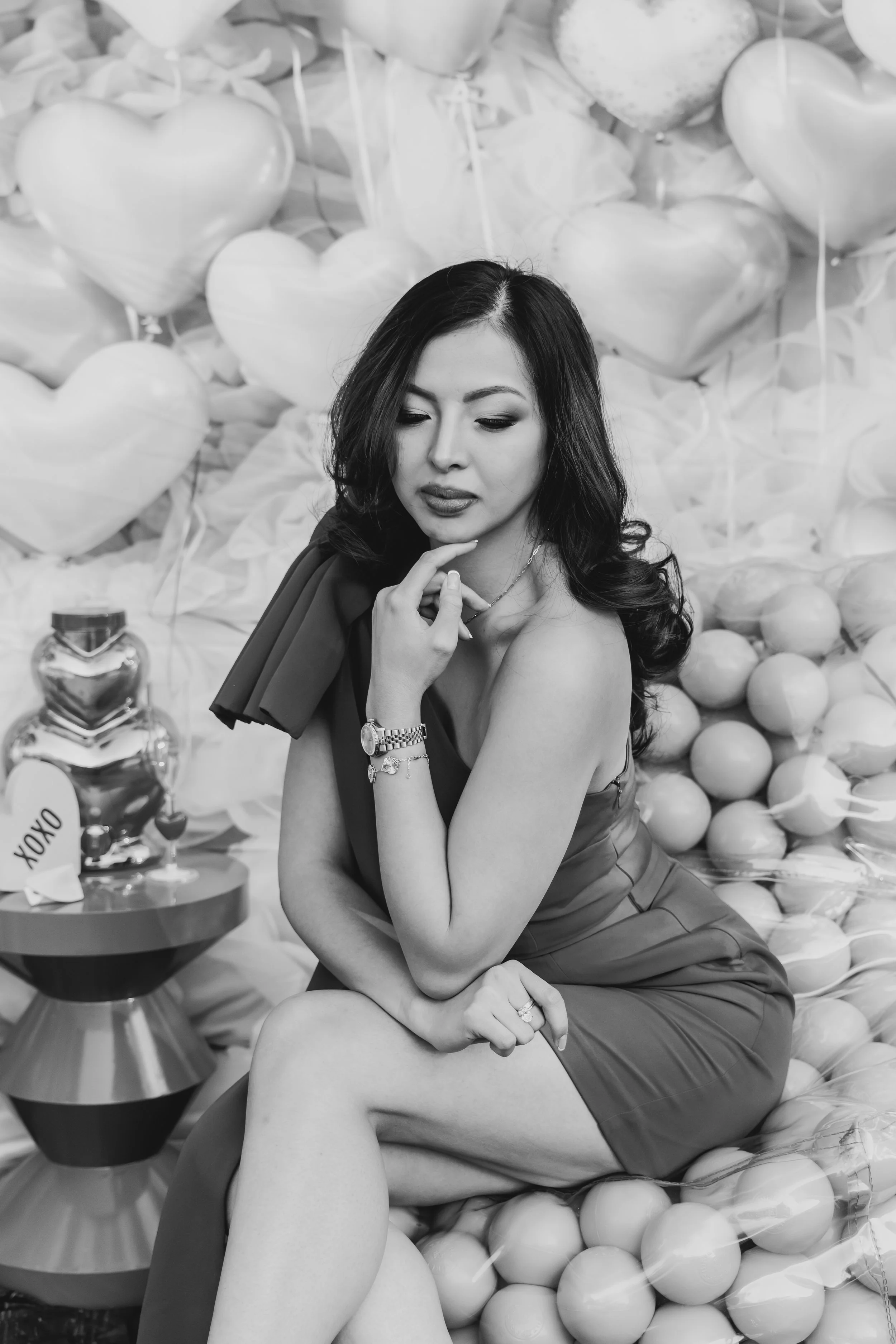 A woman in a dress sitting among balloons, with her hand on her chin, in front of a background with more balloons and gift boxes.
