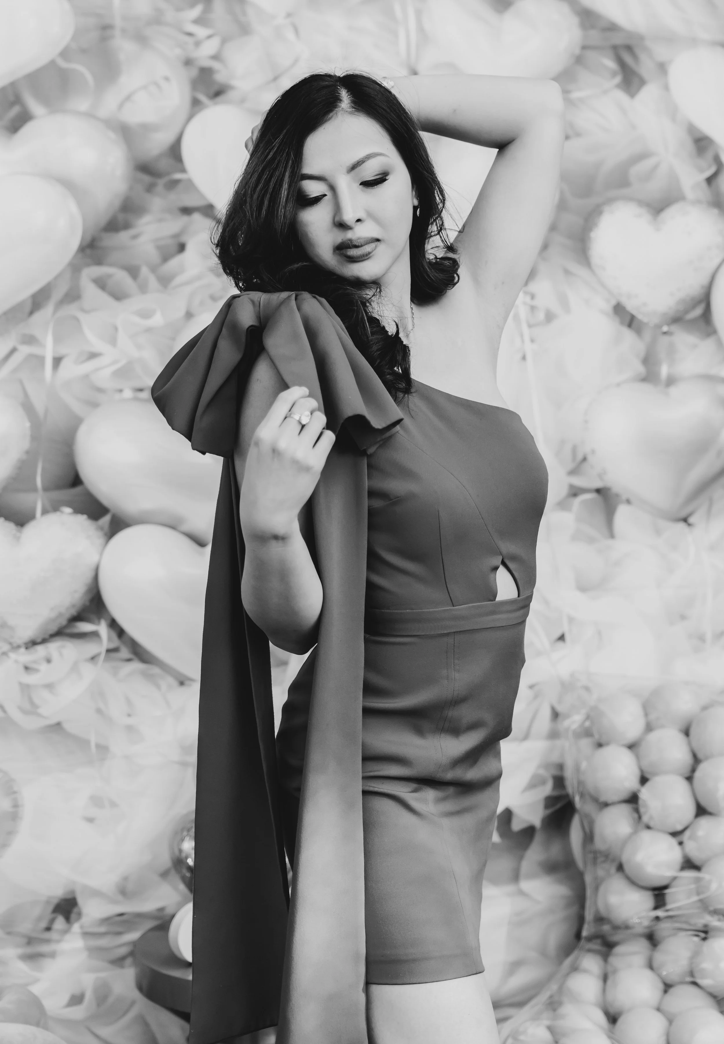 A woman with dark hair in a strapless dress holding a large fabric over her shoulder. She has her eyes closed and one hand behind her head, standing in front of a background decorated with heart-shaped and round balloons.