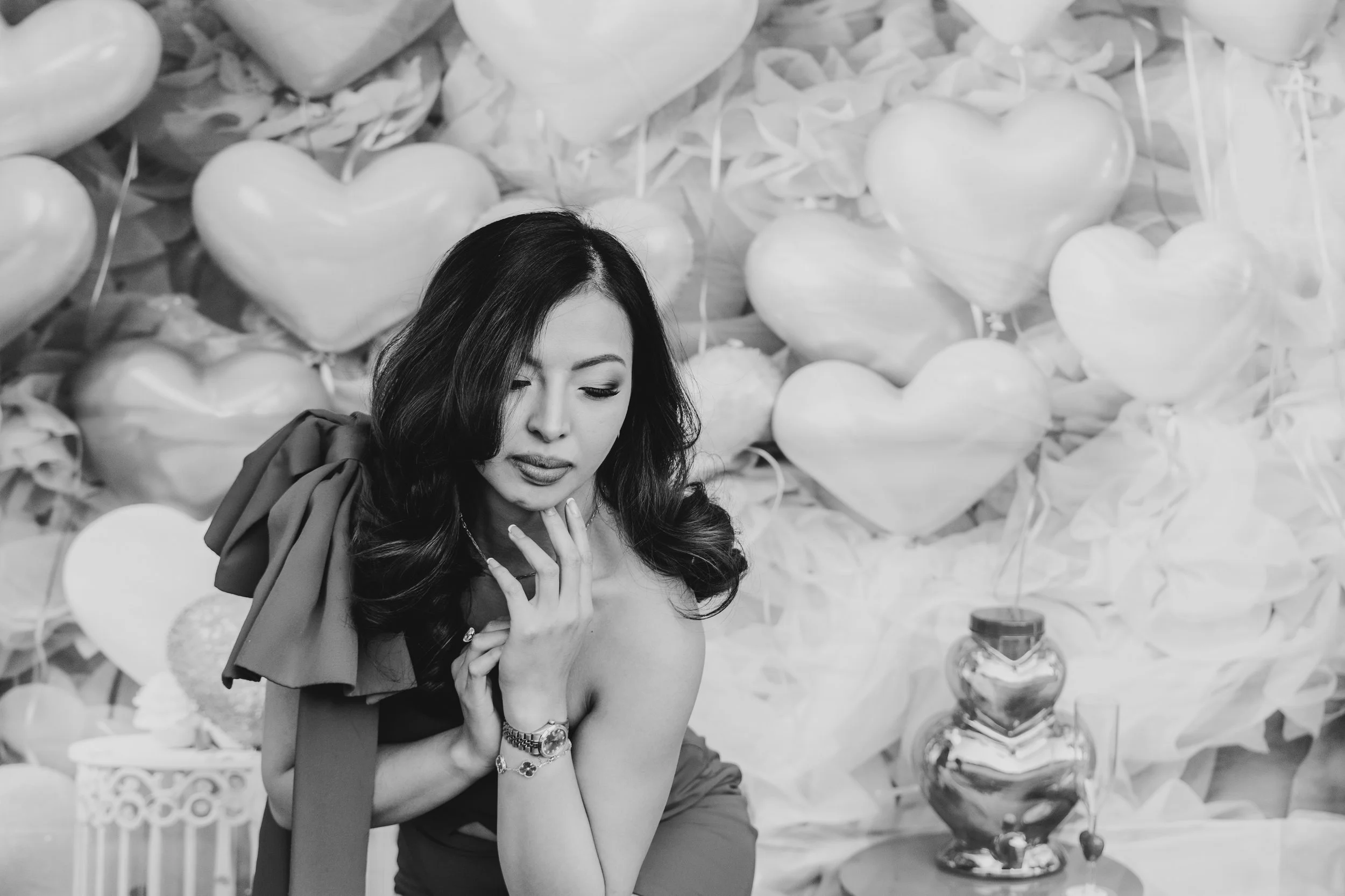 A woman with long wavy hair styled to one side, wearing a dress and jewelry, poses in front of a backdrop decorated with heart-shaped balloons and other decorative elements in a black-and-white photograph.