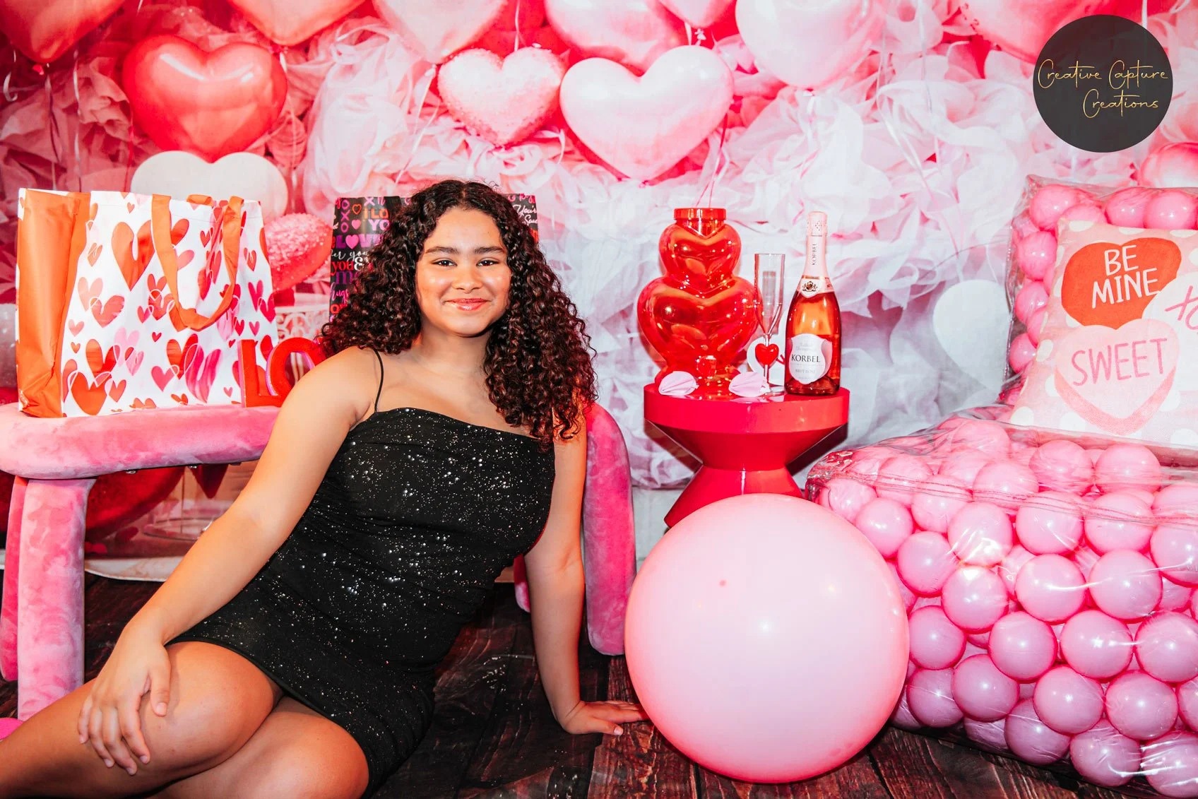 A young woman with curly hair wearing a black dress sitting on the floor in front of a Valentine's Day themed backdrop, decorated with pink hearts, balloons, and Valentine's themed pillows that say "Be Mine" and "Sweet."