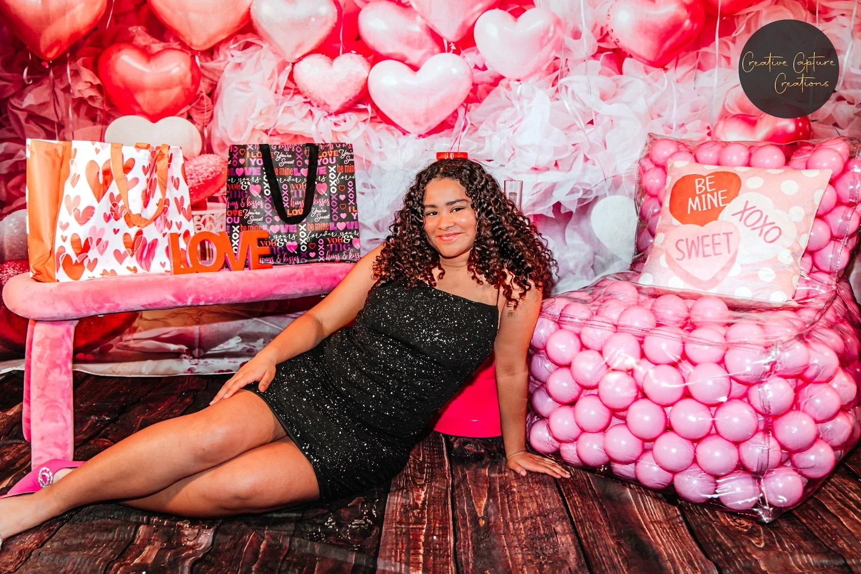 A woman with curly hair in a black, sparkly dress is lying on the floor, leaning on her left arm. Behind her is a Valentine's Day-themed backdrop with pink and red balloons, hearts, and love-themed decorations. To her right is a pink armchair filled 