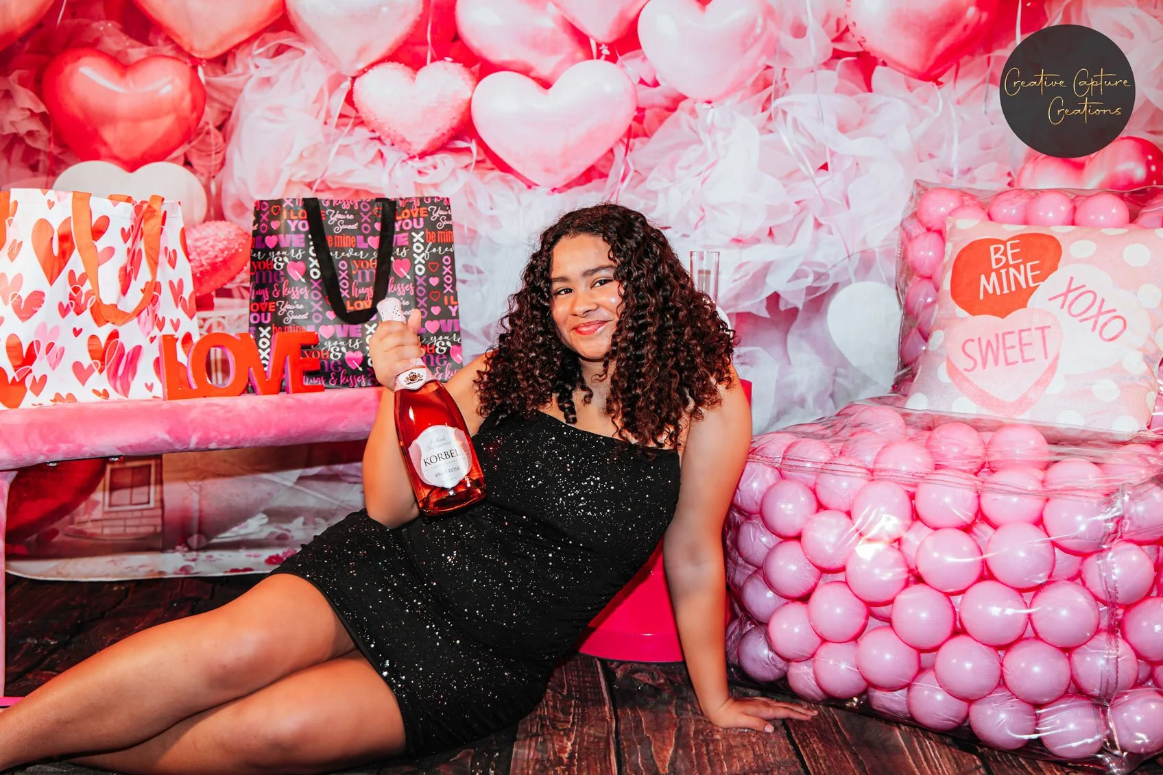 A woman with curly hair in a black sparkly dress is sitting on the floor, holding a bottle of pink wine, with a Valentines Day themed backdrop of pink and red hearts, gift bags, and a pink ball pit.
