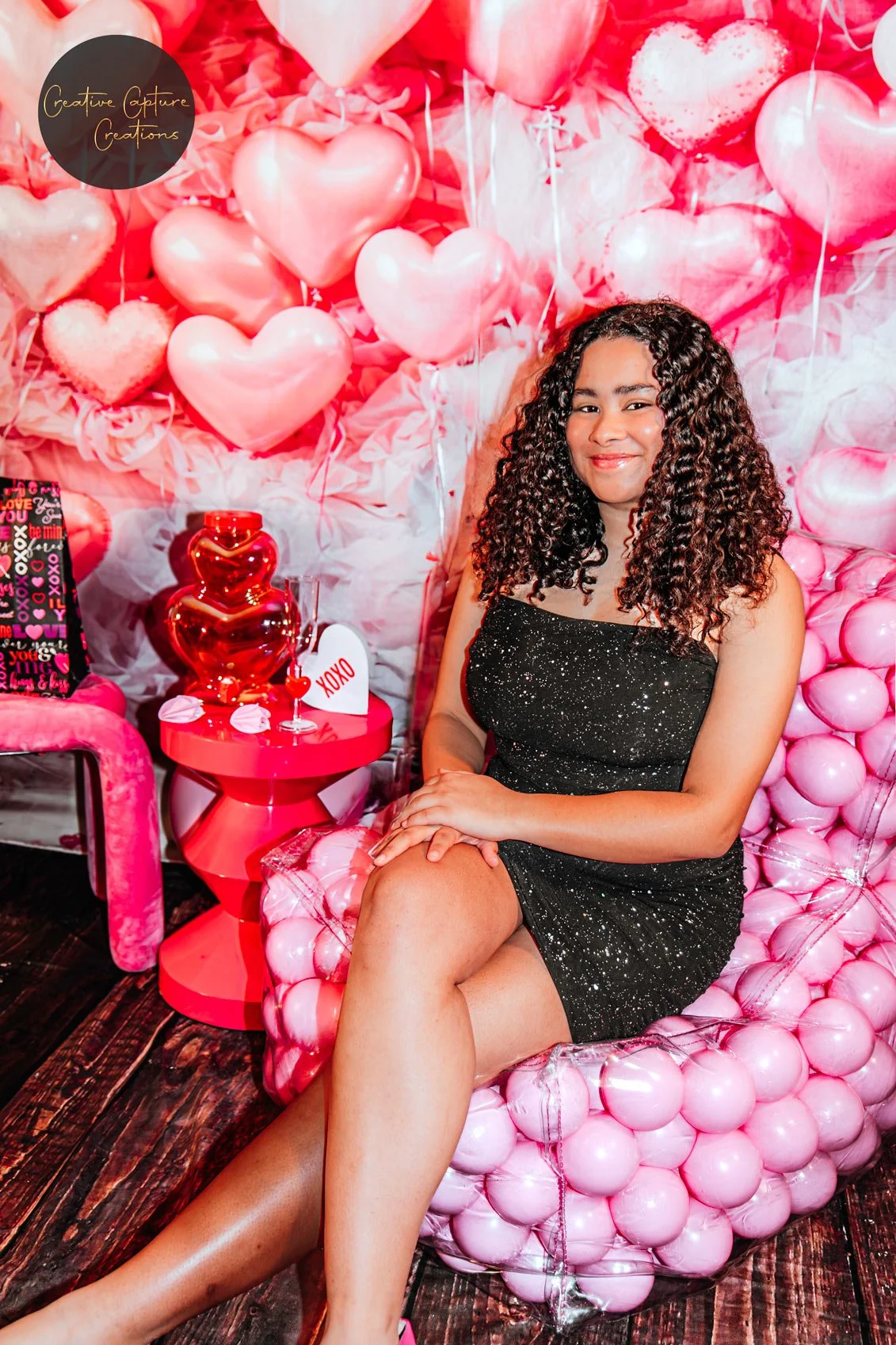 A young woman with curly hair sitting on a pink balloon chair with a Valentine's Day theme background of pink and red balloons, featuring hearts and love-themed decorations.