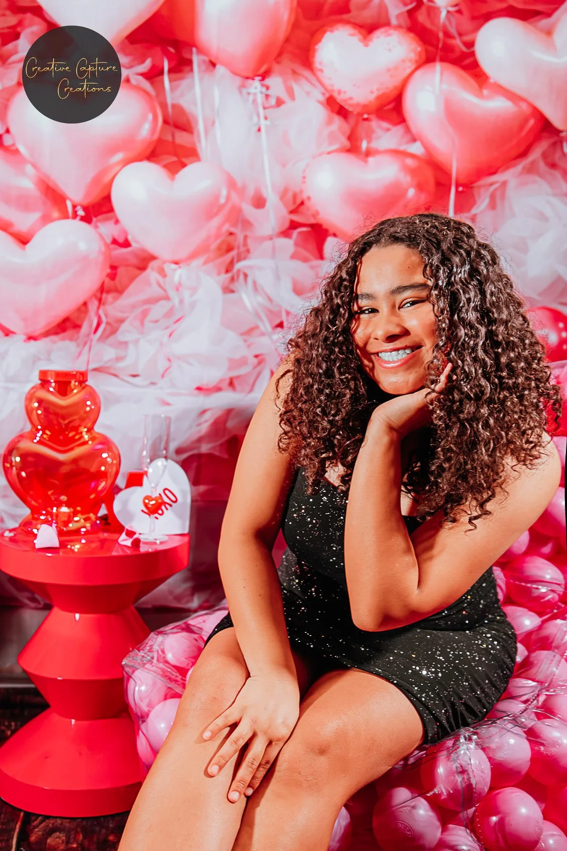 A woman with curly hair smiling and sitting on a pink balloon and fabric backdrop decorated with heart-shaped balloons in shades of pink and red. There is a red table with a red bear-shaped ornament and a tag that reads "LOVE".