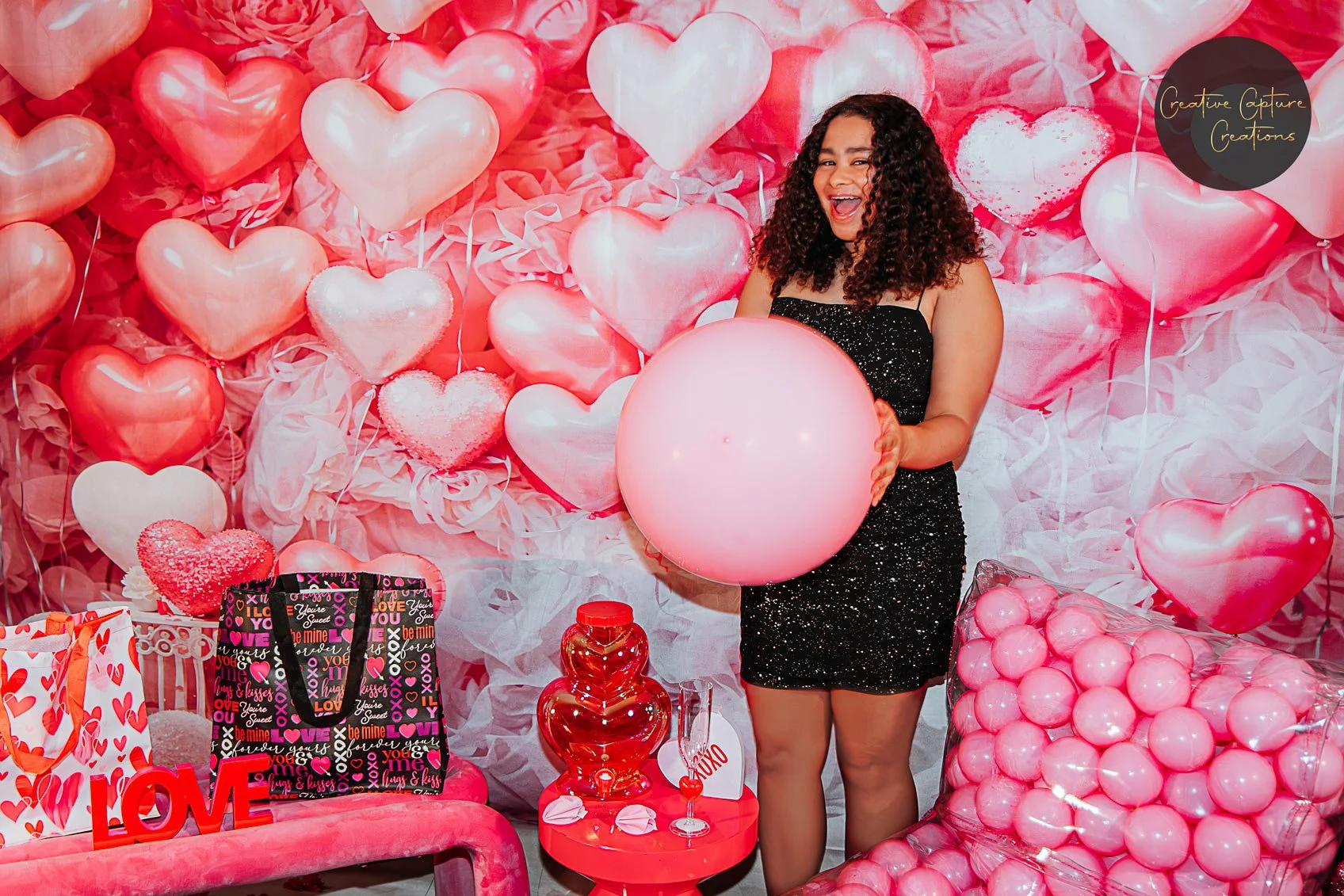 A woman with curly hair in a black sparkly dress holding a large pink balloon, smiling at a Valentines Day themed party with pink and red heart decorations, balloons, gift bags, and a red glass lamp.