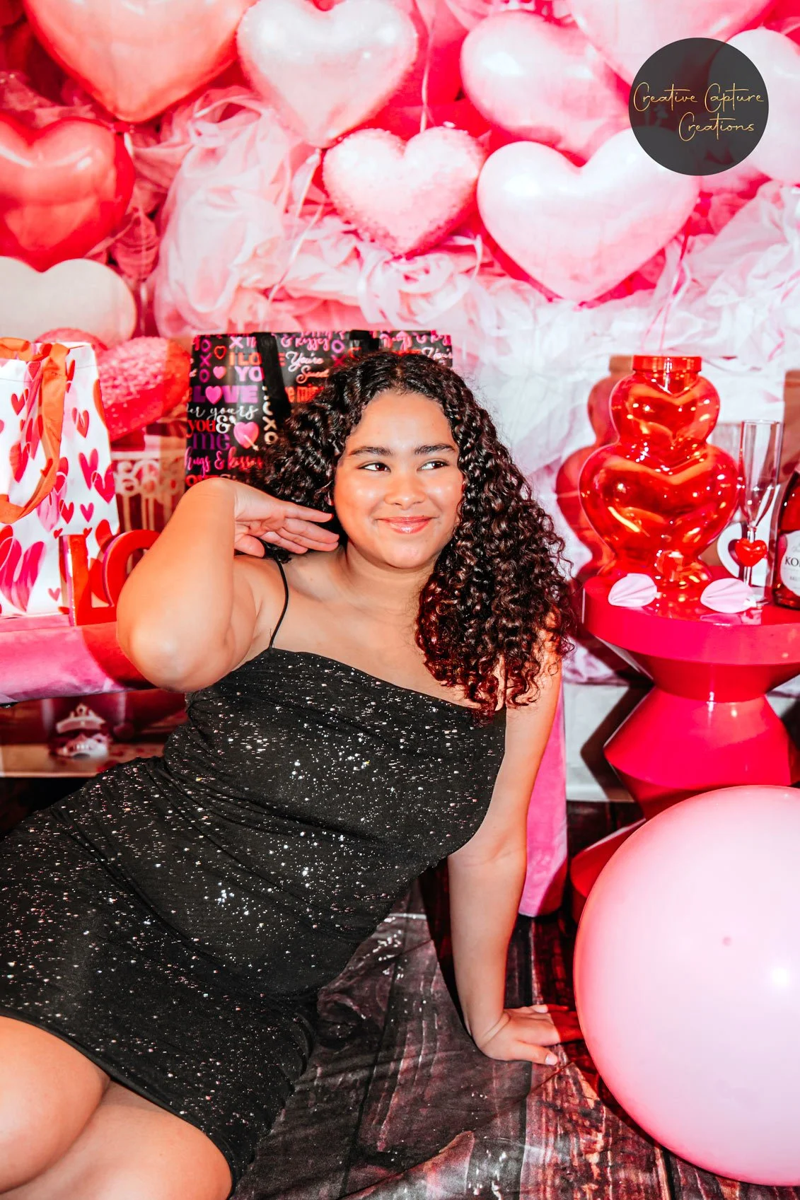 A young woman with curly hair and a black sparkly dress sitting on the floor, smiling, in front of a Valentine's Day themed backdrop with pink and red balloons, gift bags, and decorative hearts.