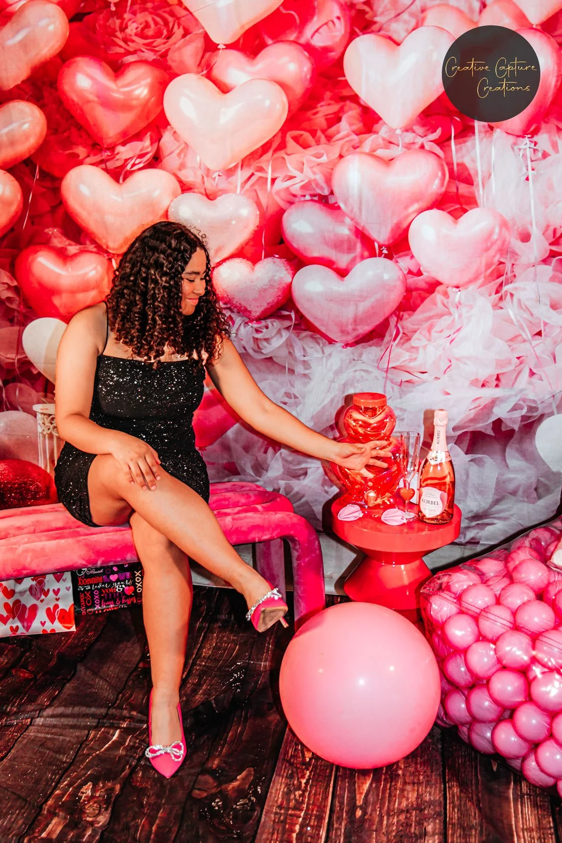 A woman with curly hair dressed in a black sparkly dress and pink high heels sitting on a pink bench, touching a large red heart-shaped vessel. The background features a wallpaper of various pink and red heart balloons and paper decorations, with a s