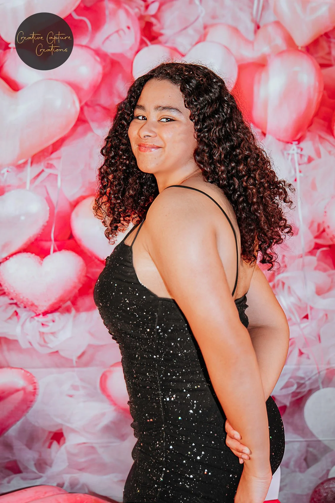 Young woman with curly hair wearing a black, star-spangled dress, smiling with a pink heart-themed backdrop.