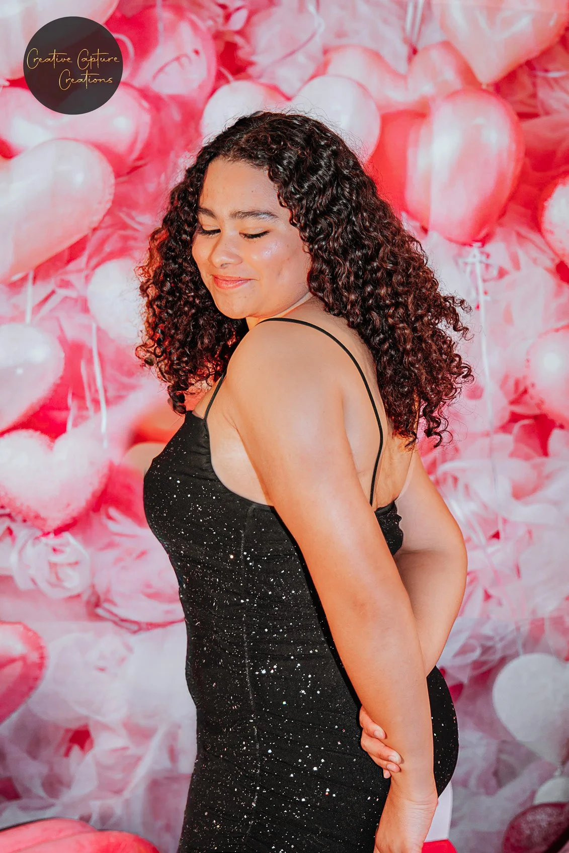 A woman with curly hair wearing a black, sparkly dress with spaghetti straps, standing in front of a backdrop of pink and white heart-shaped balloons.