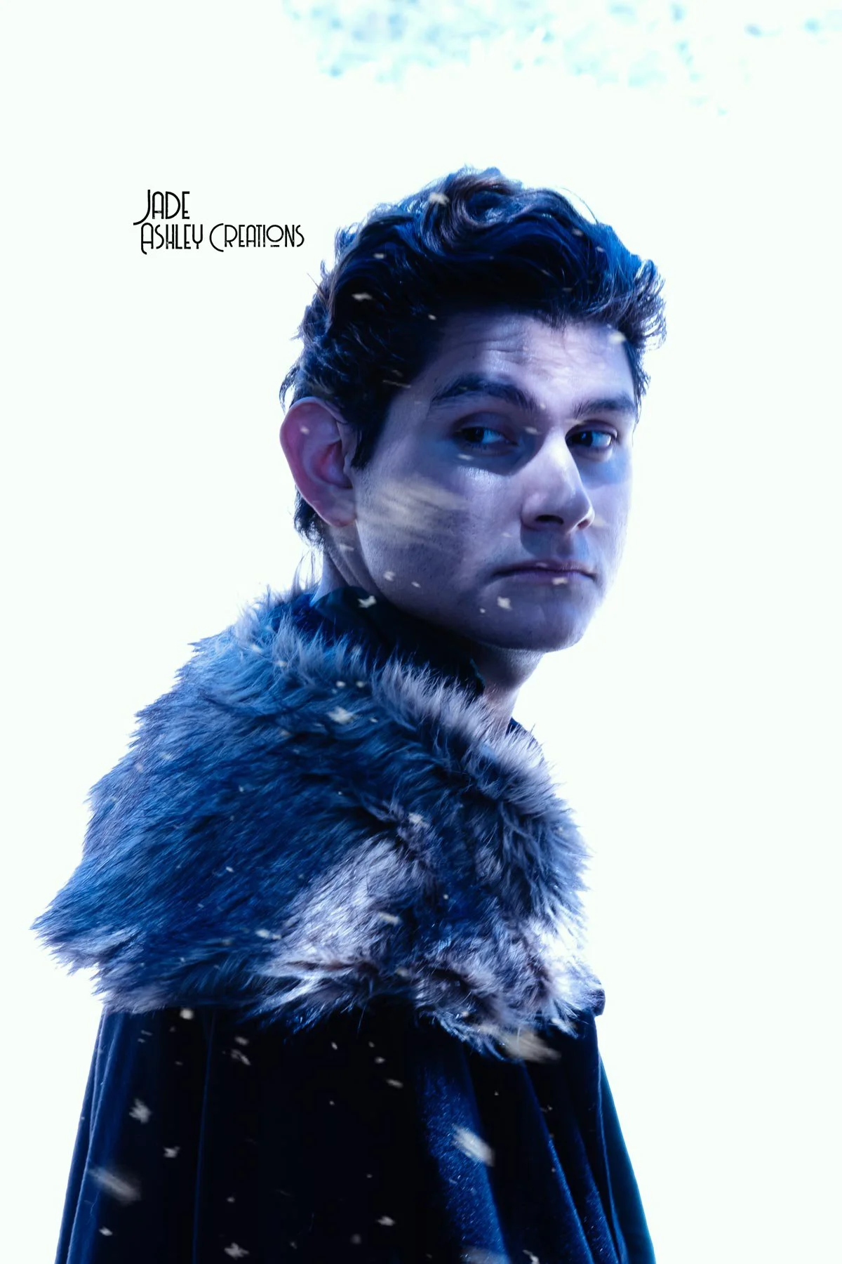 A young man with dark wavy hair wearing a fur-lined cloak outdoors in a snowy environment with snowflakes falling.