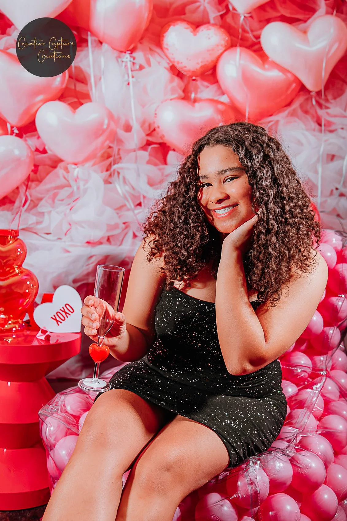A young woman with curly hair sitting on balloons and decorations with a Valentine's Day theme, holding a glass and smiling.