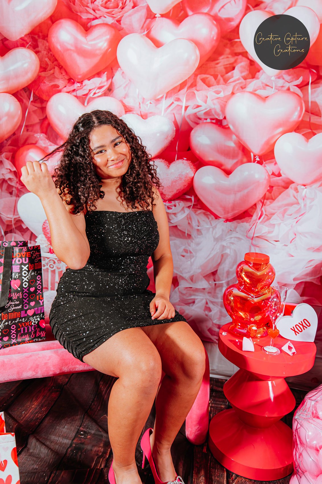 A woman sitting on a pink velvet bench in front of a Valentine-themed backdrop with pink and white heart-shaped balloons and pink tissue paper. She is smiling, wearing a black dress with sparkles and pink high heels. There are Valentine's Day decorat